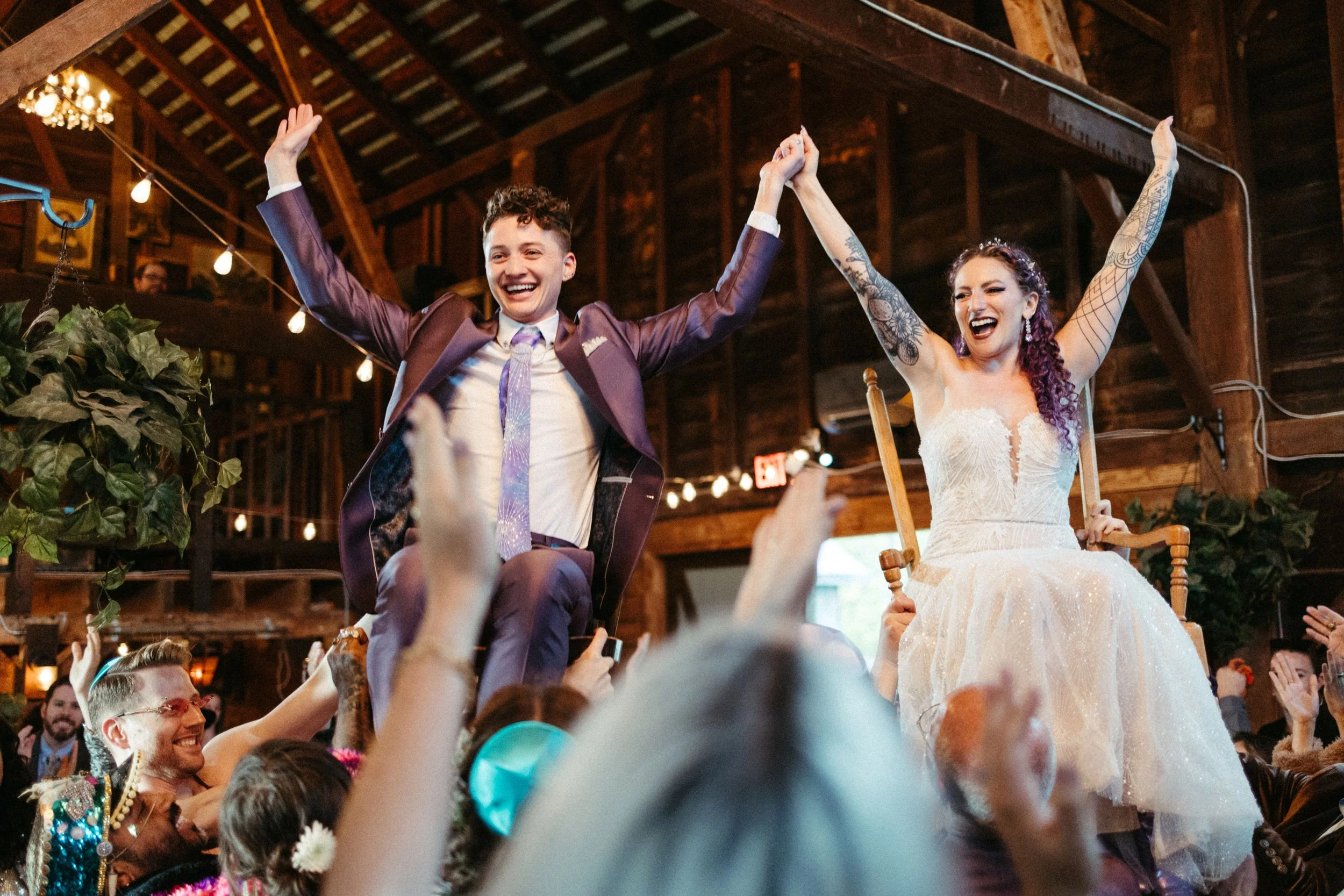 A bride and groom sitting on chairs are lifted by guests during a celebration, with the bride wearing a white wedding dress and the groom in a purple suit, inside a rustic wooden venue with string lights.