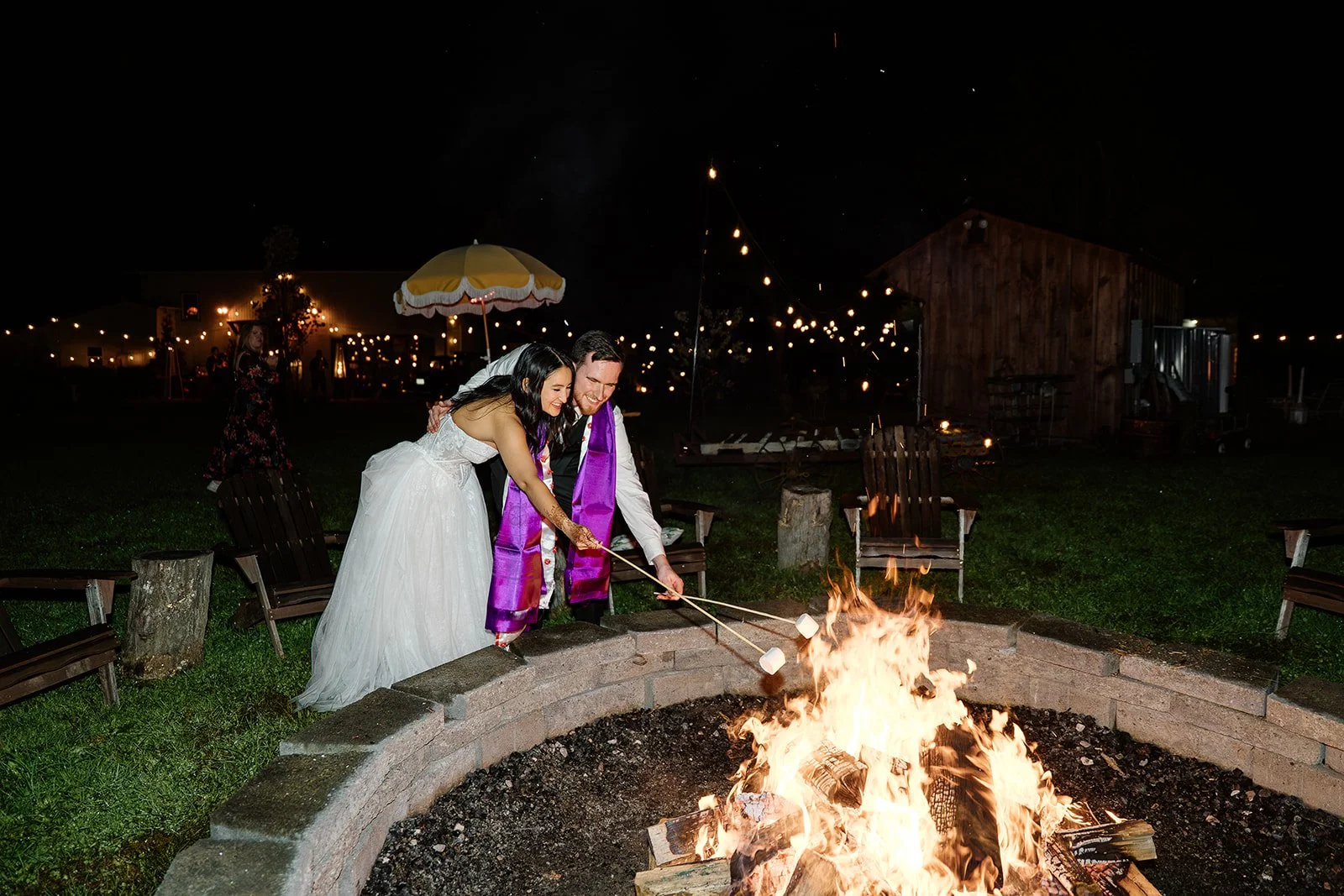 A bride and groom roasting marshmallows over a fire pit at night during their wedding celebration, with string lights and a wooden building in the background.