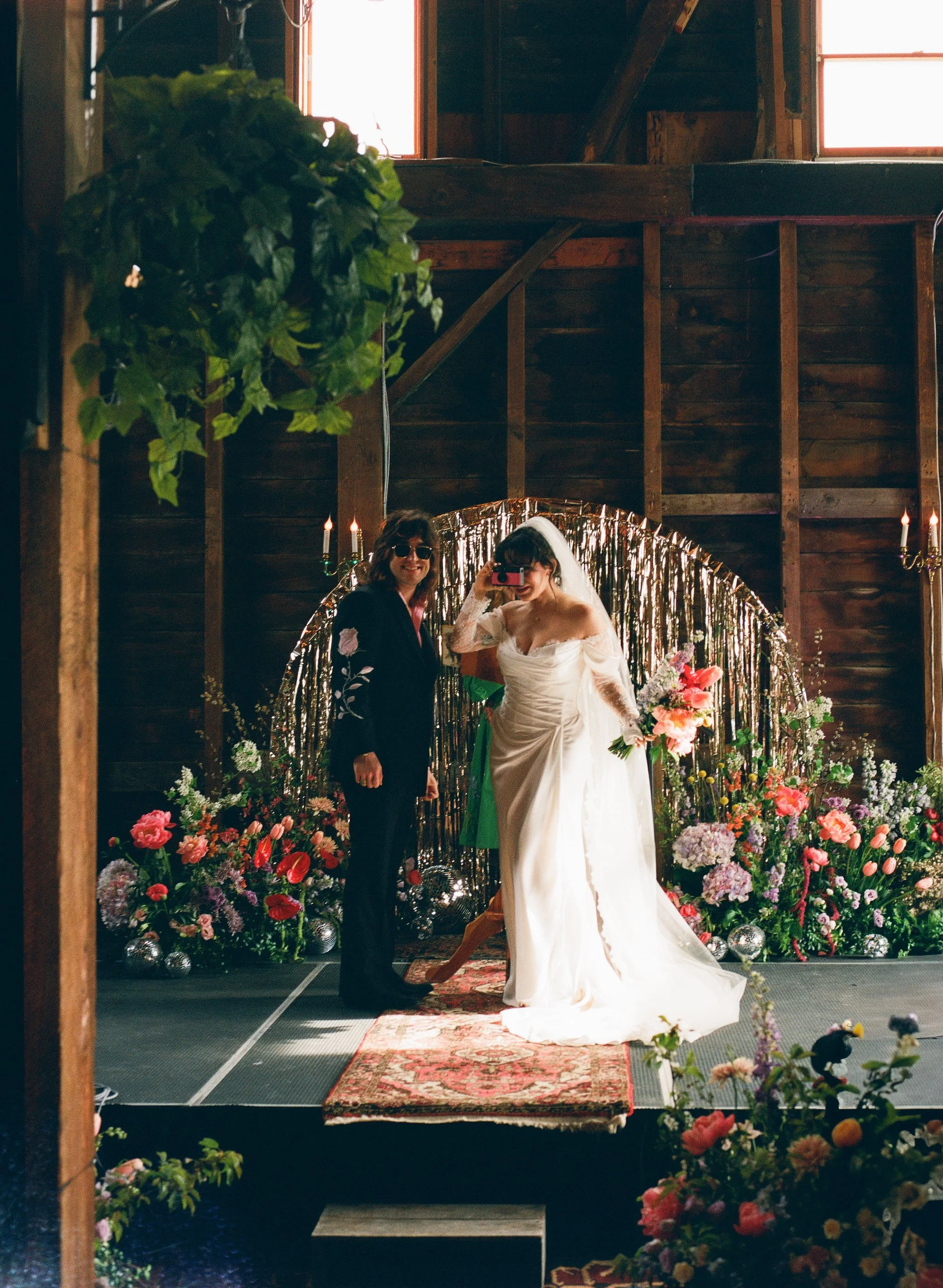 Wedding ceremony with a bride in a white gown and veil, standing next to a woman in dark clothing, in front of a decorative floral backdrop inside a rustic wooden barn.