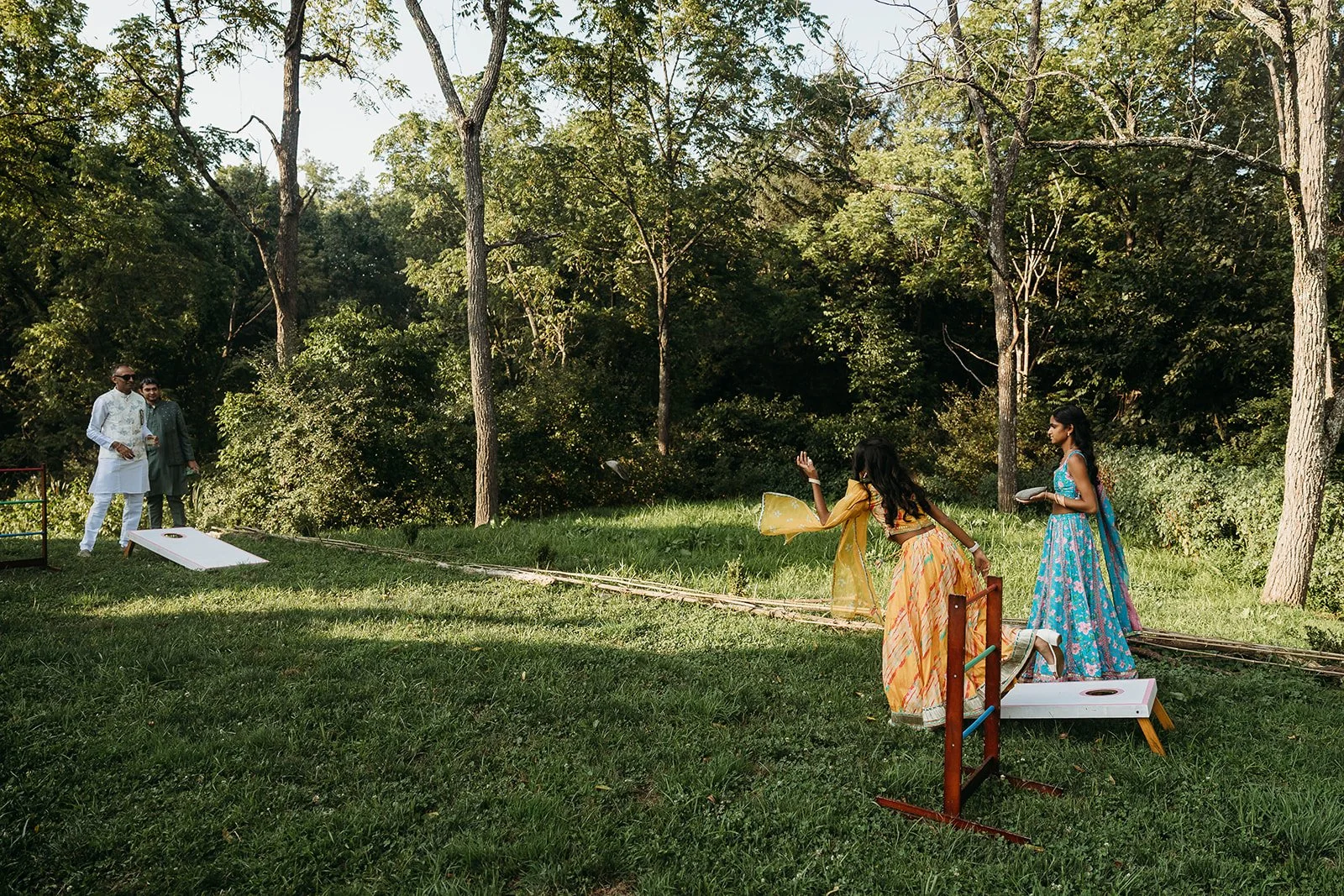 People playing corn hole game outdoors in a wooded area, dressed in traditional Indian clothing.