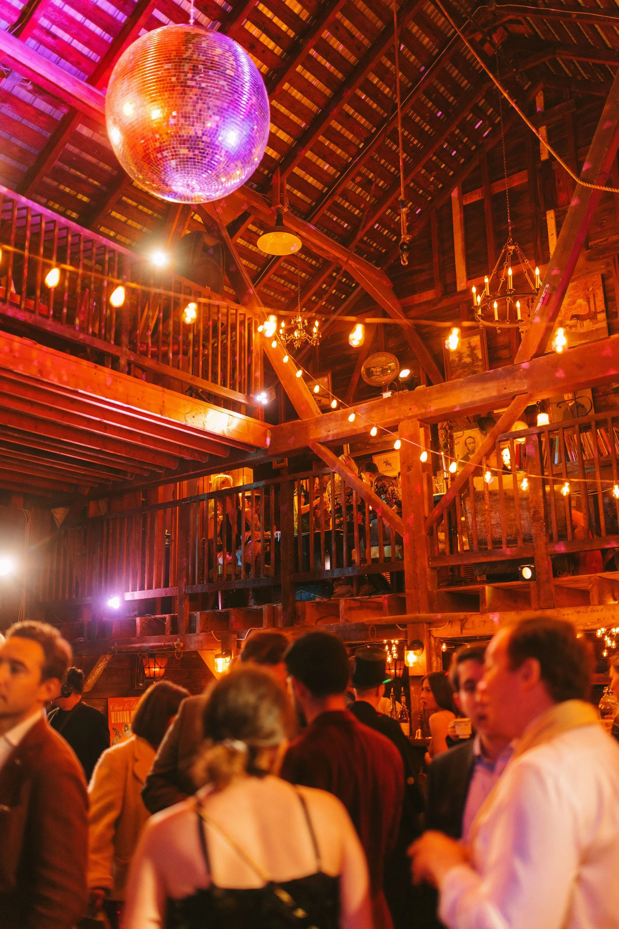Interior of a lively bar or restaurant with wooden beams, string lights, and people socializing, illuminated by warm and colorful lighting.