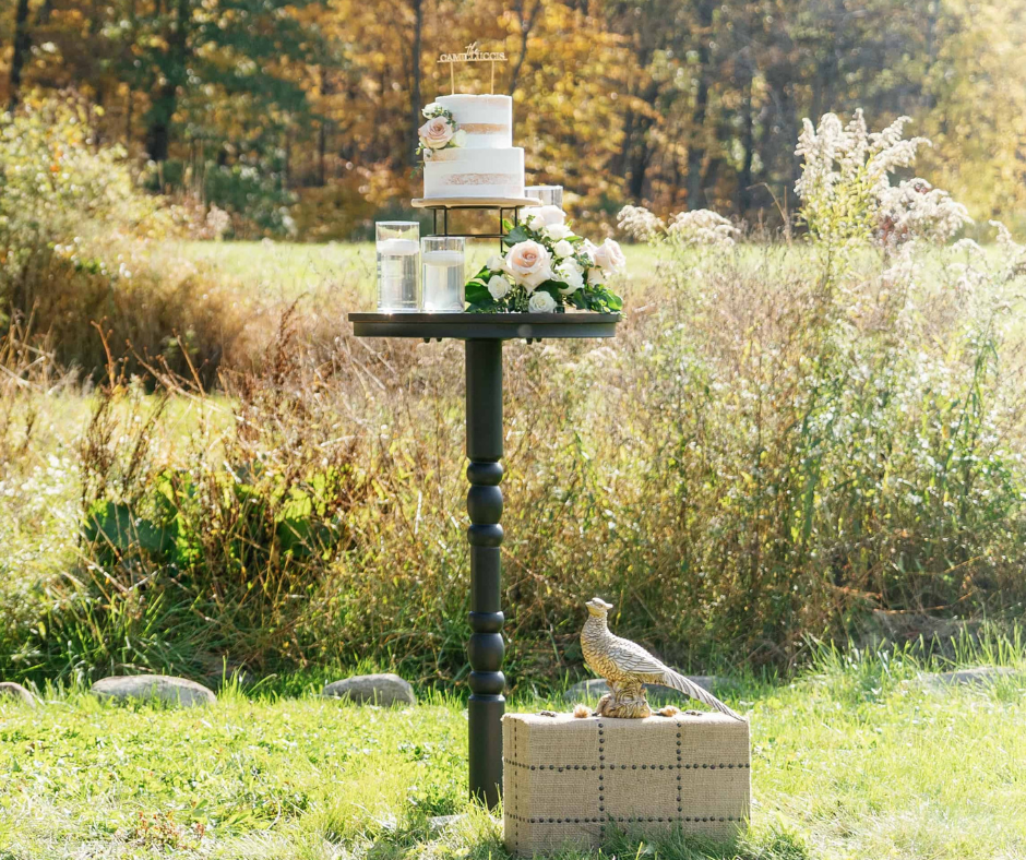 A wedding cake with flowers on top, placed on a table outdoors in a natural setting, with rocks, grass, and plants surrounding it, and a decorative hawk statue on a cushion at the base.