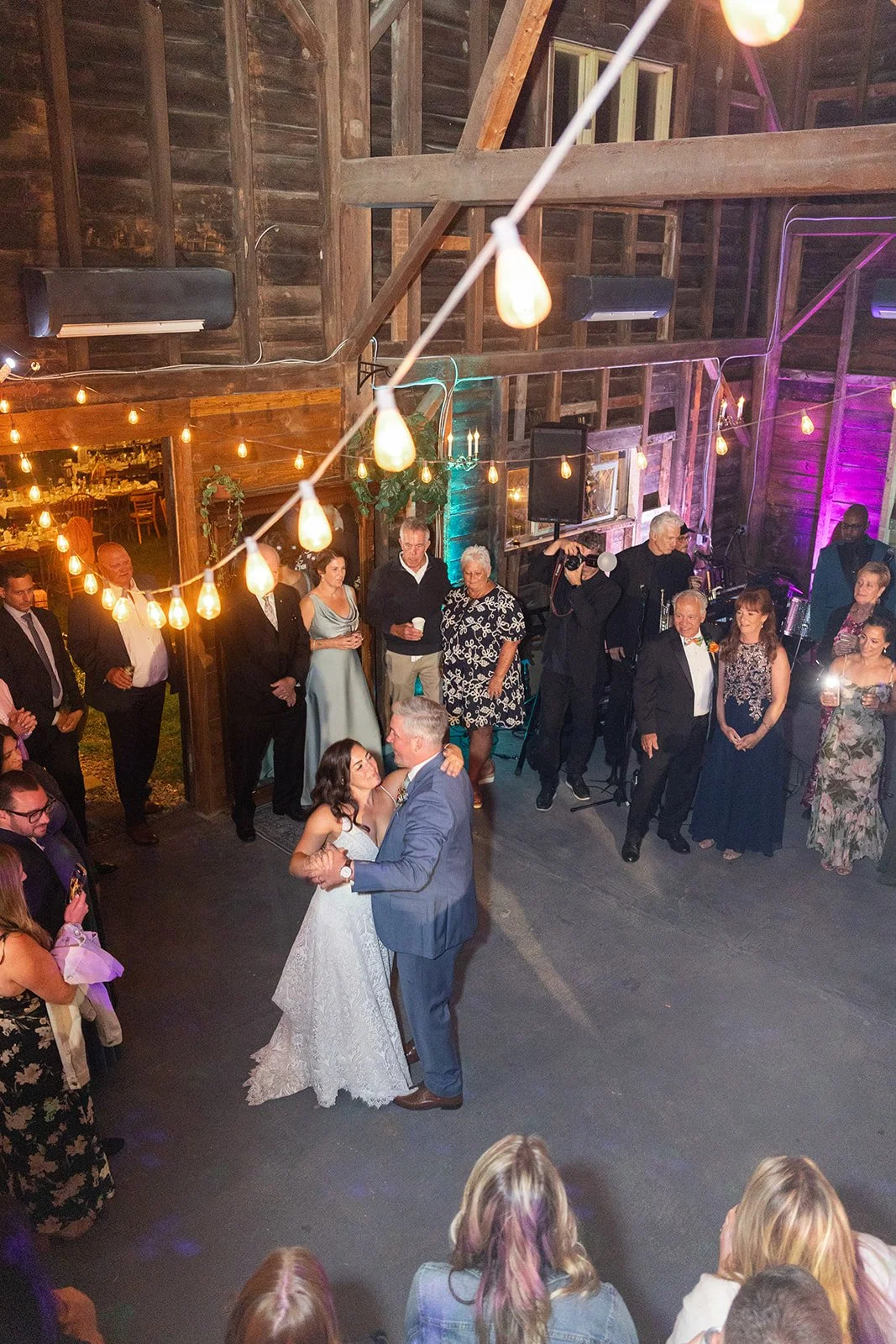 A bride and groom share a dance inside a rustic barn decorated with string lights and guests watching, during a wedding reception.