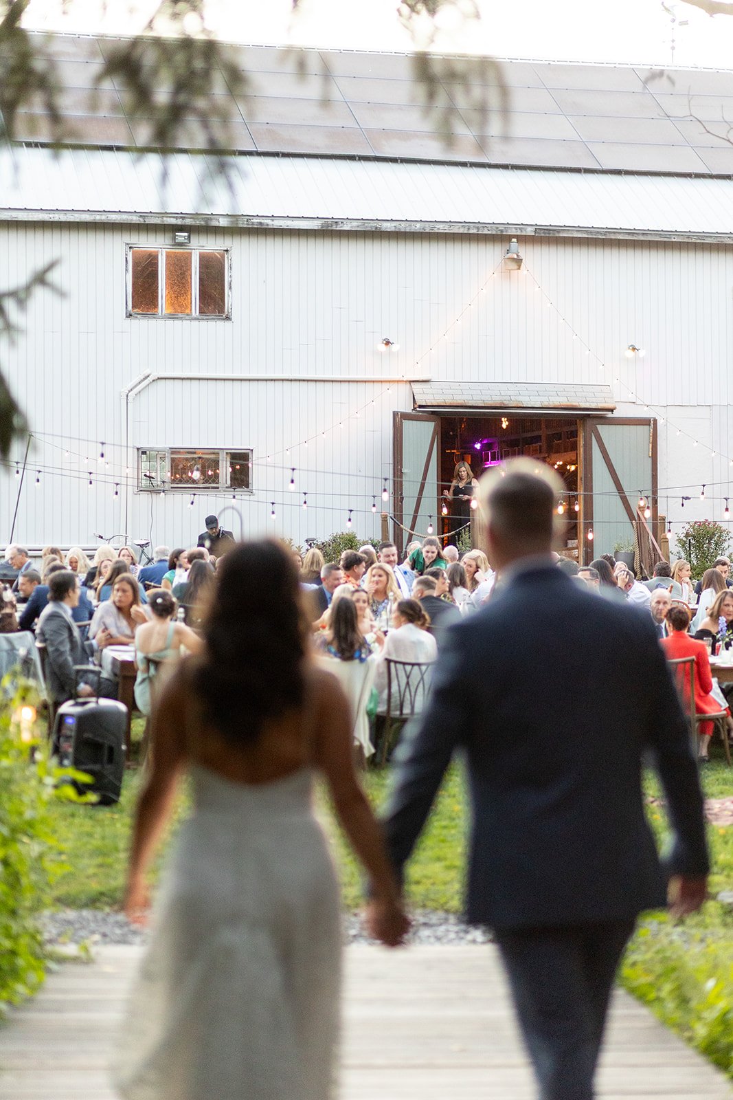 A bride and groom walking hand in hand towards a wedding reception with guests seated at tables outside a barn decorated with string lights.