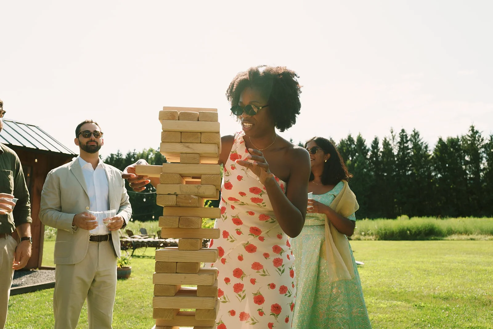 A group of people outdoors playing giant Jenga, a woman in a floral dress is about to remove a block from the game, others watch and smile.