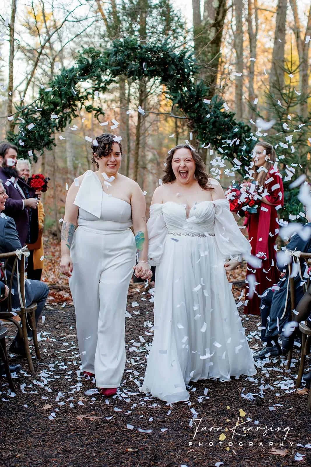 Two women, one in a white strapless gown and the other in a white jumpsuit, holding hands and smiling, walking through falling confetti at an outdoor wedding ceremony amid autumn trees.