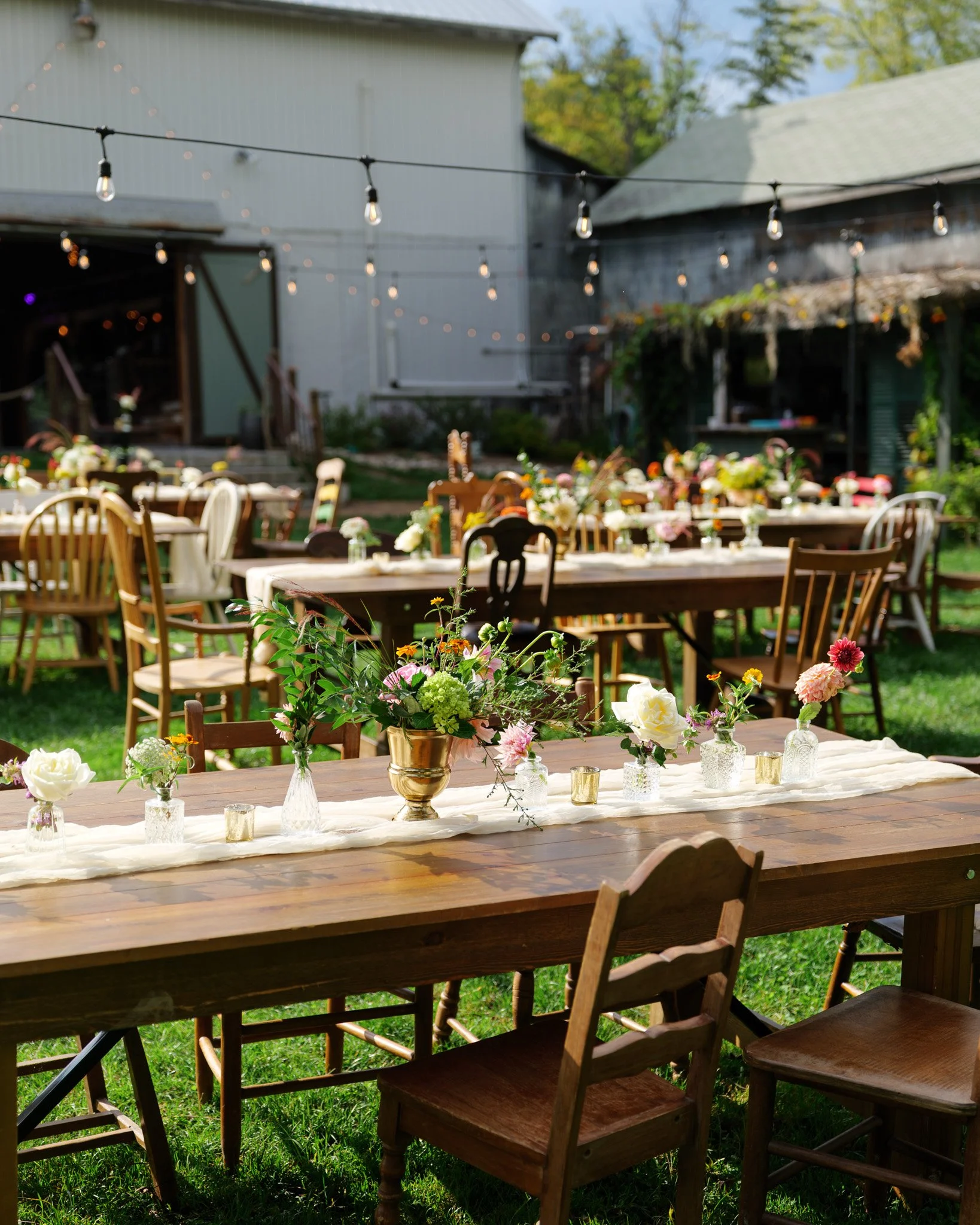 Outdoor garden party setup with wooden tables and chairs, decorated with flowers in vases and string lights