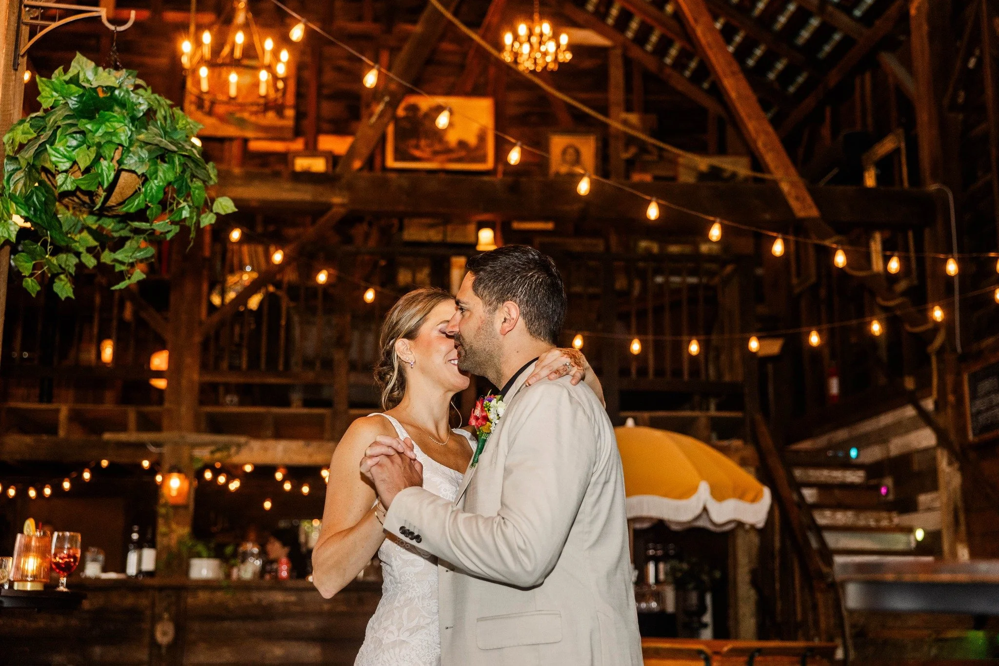 A bride and groom dance closely in a rustic barn decorated with string lights and warm ambient lighting.