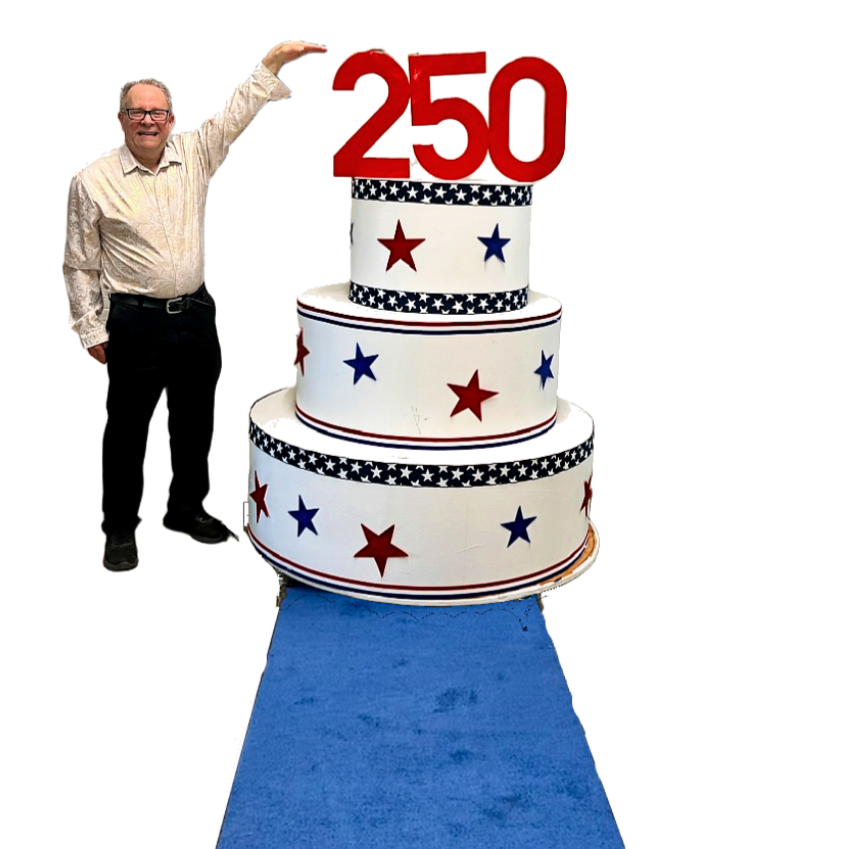 A man standing next to a large, three-tiered cake decorated with red, white, and blue stars and stripes, with a red "250" topper, on a blue carpet.