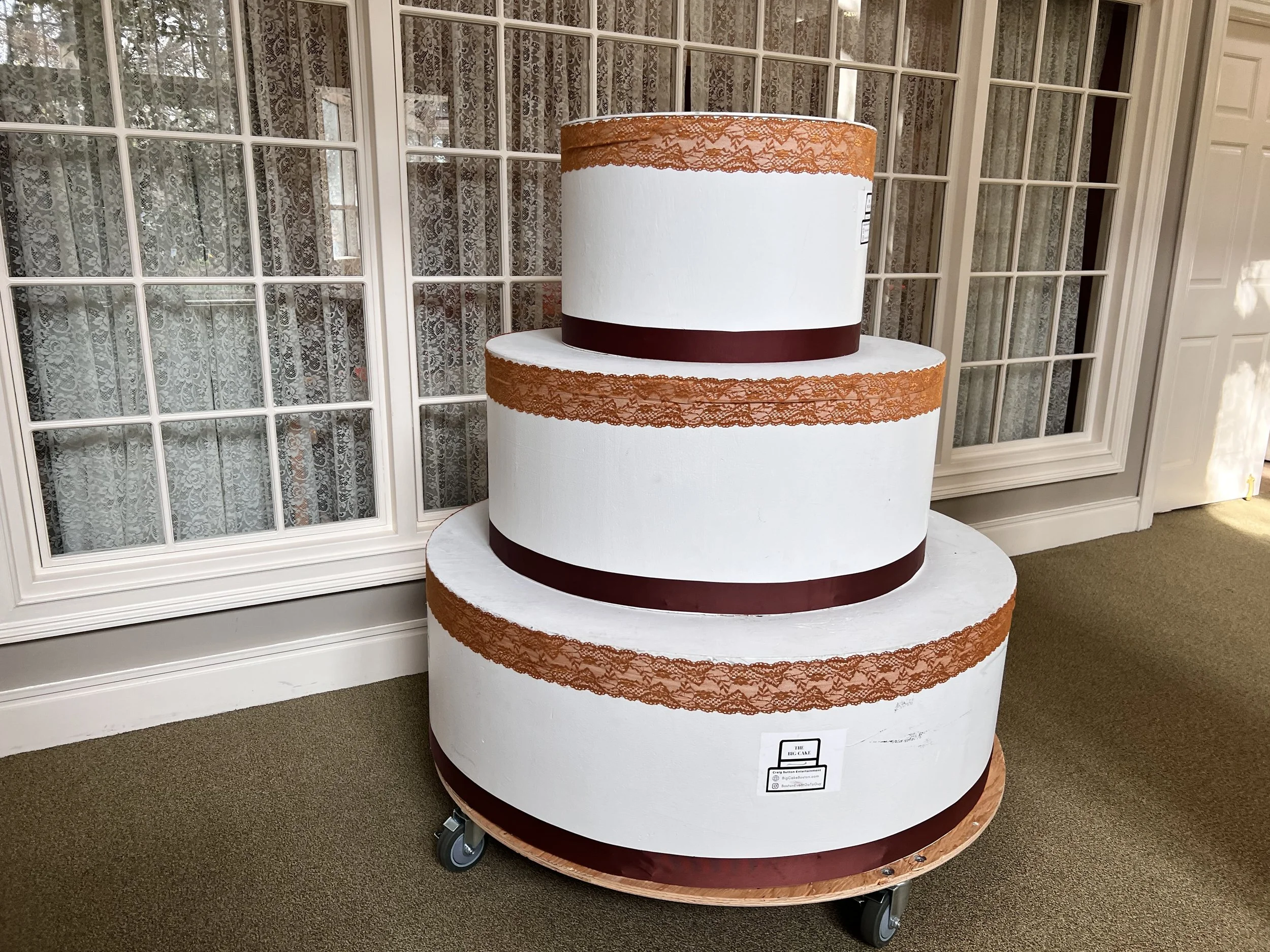 Three-tiered white wedding cake with rust-colored lace and dark red bands on each tier, positioned on a wheeled wooden base in front of lanai windows with lace curtains.