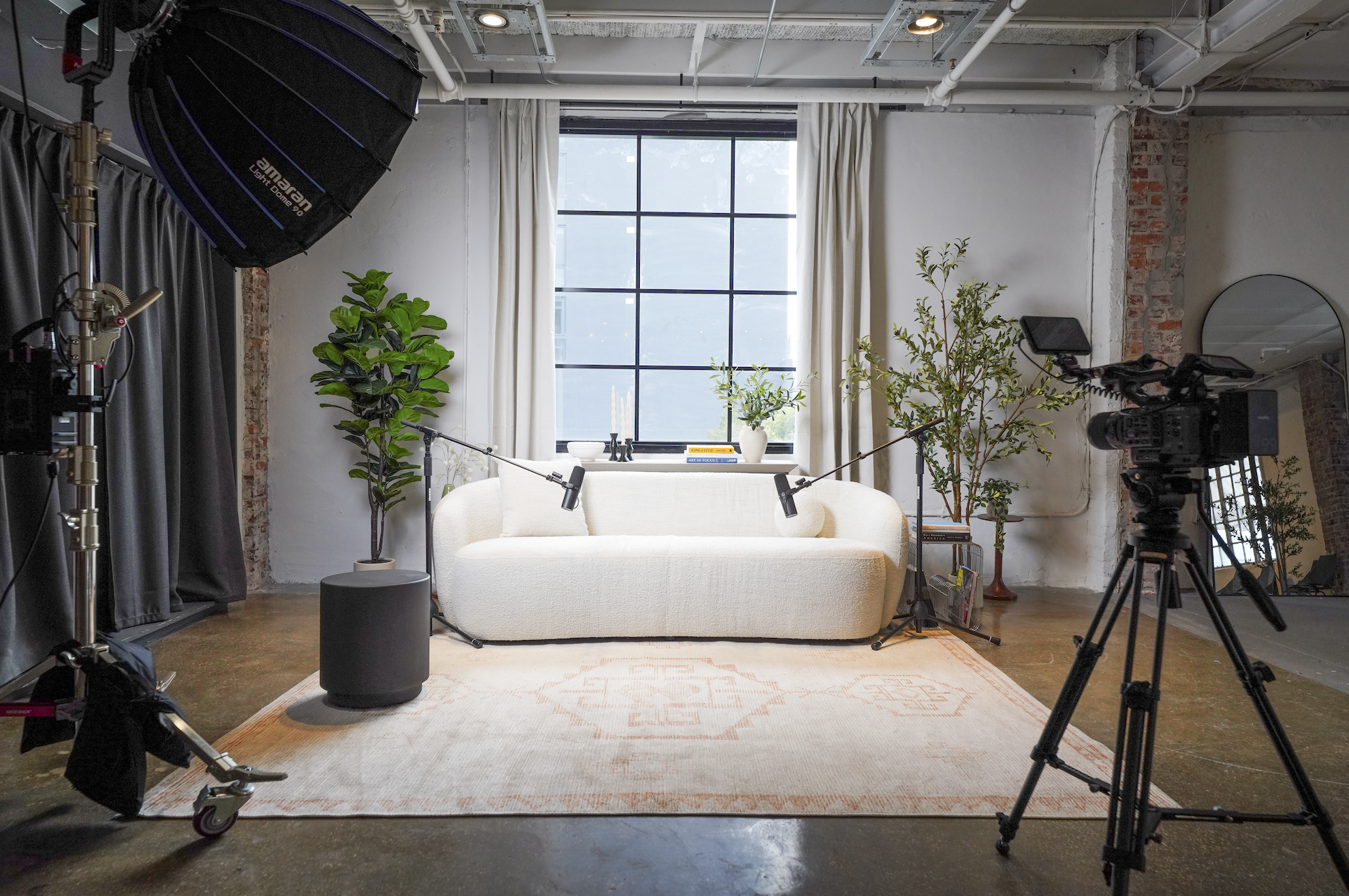 Two people sitting in a room with beige curtains and potted plants in the background, posing for a photo. The woman on the left is wearing a tan plaid blazer, white shirt, and jeans. The man on the right is wearing a green button-up shirt, khaki pants, and sneakers, with one leg crossed over the other.