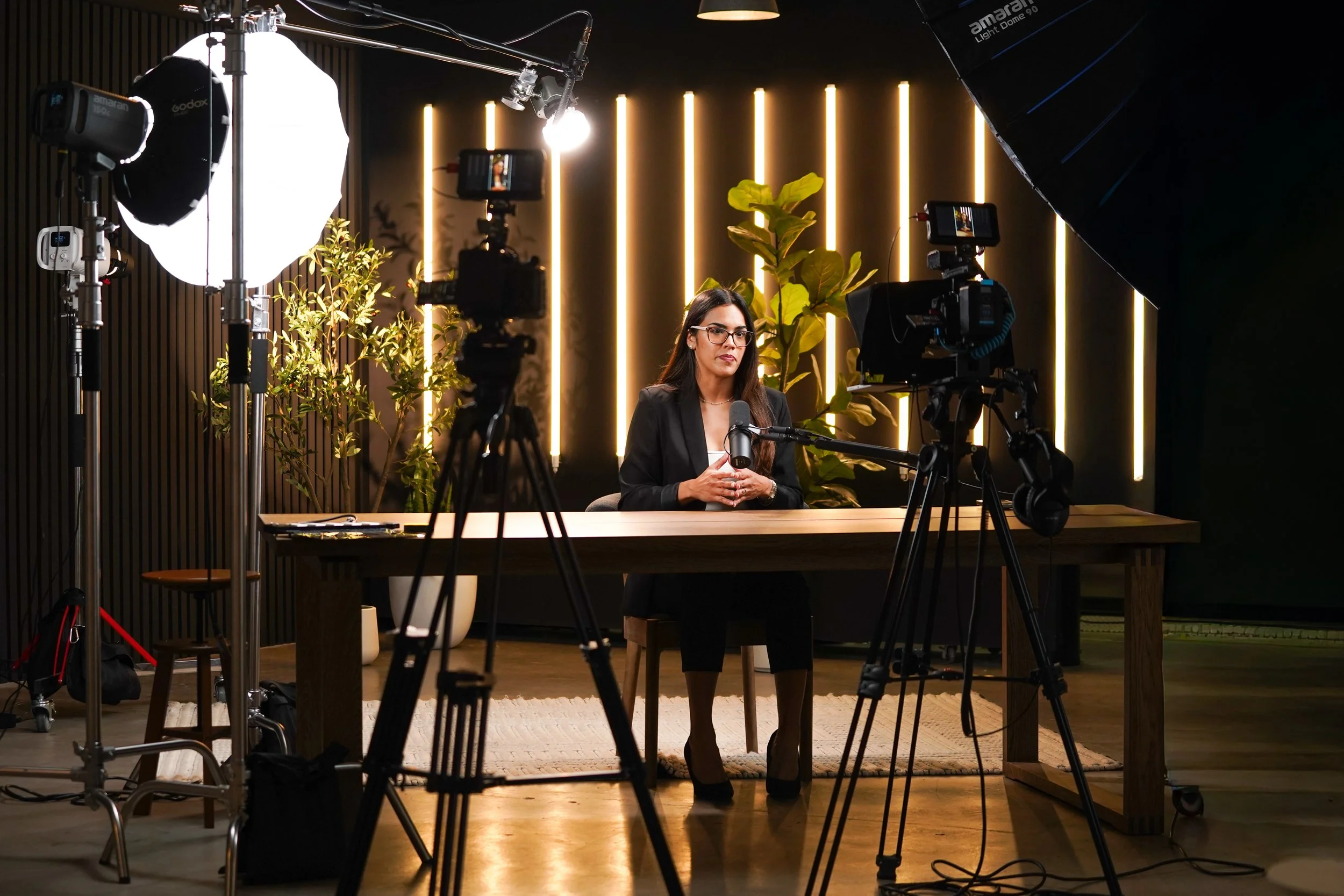 A woman and a man having a podcast conversation, sitting in armchairs with a small wooden side table between them, two microphones, and plants in the background.