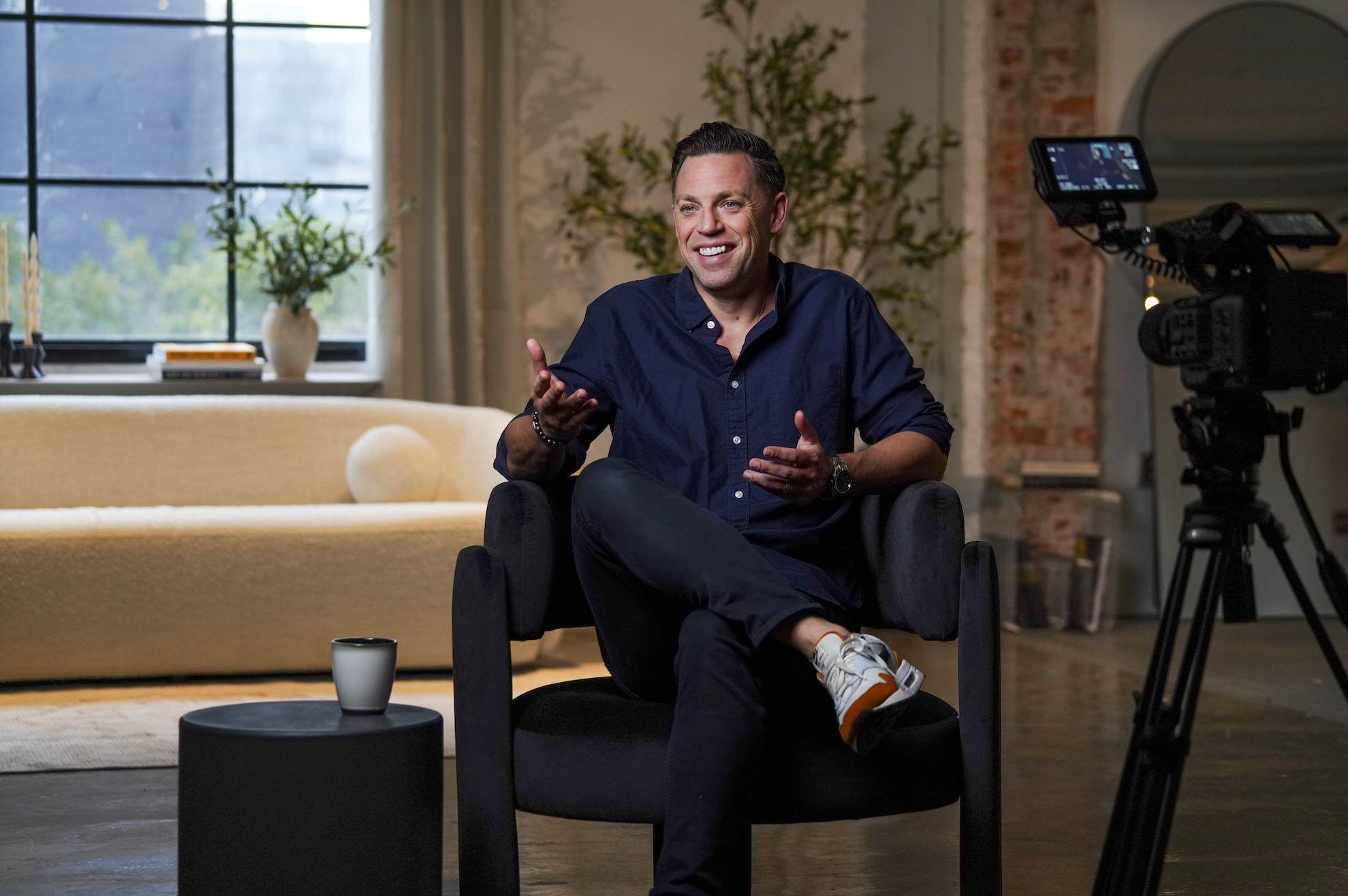Two women sitting in a living room, having a conversation with microphones in front of them, holding white mugs. The room has plants, artwork, and a window with a view outside.