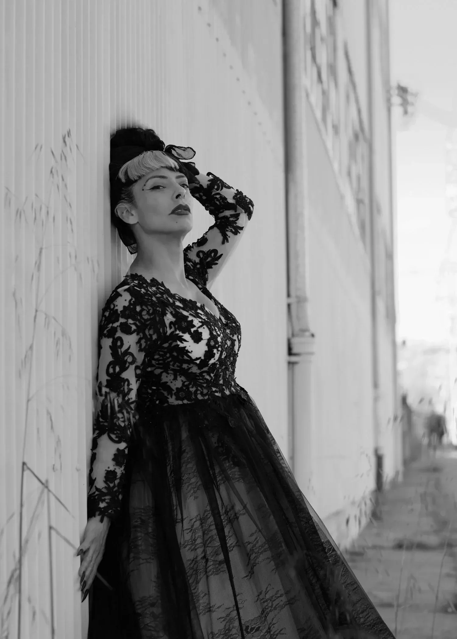 Black and white photo of a woman with dark lipstick and vintage makeup, wearing a black lace dress, leaning against a wall with a confident expression.