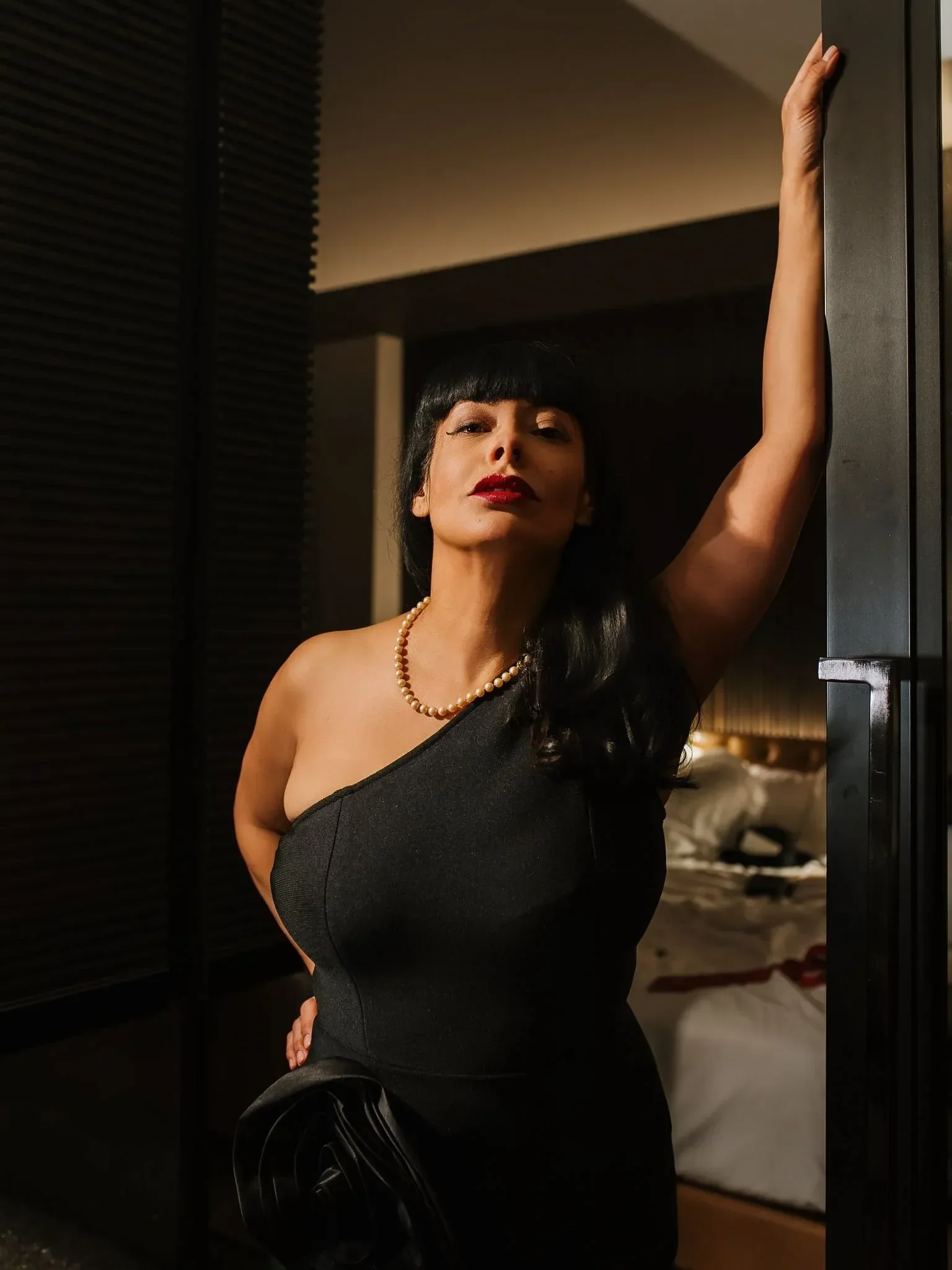 A woman with dark hair and bangs wearing a black one-shoulder dress and a pearl necklace, standing in a dimly lit room, leaning against a door frame with her arm raised above her head.
