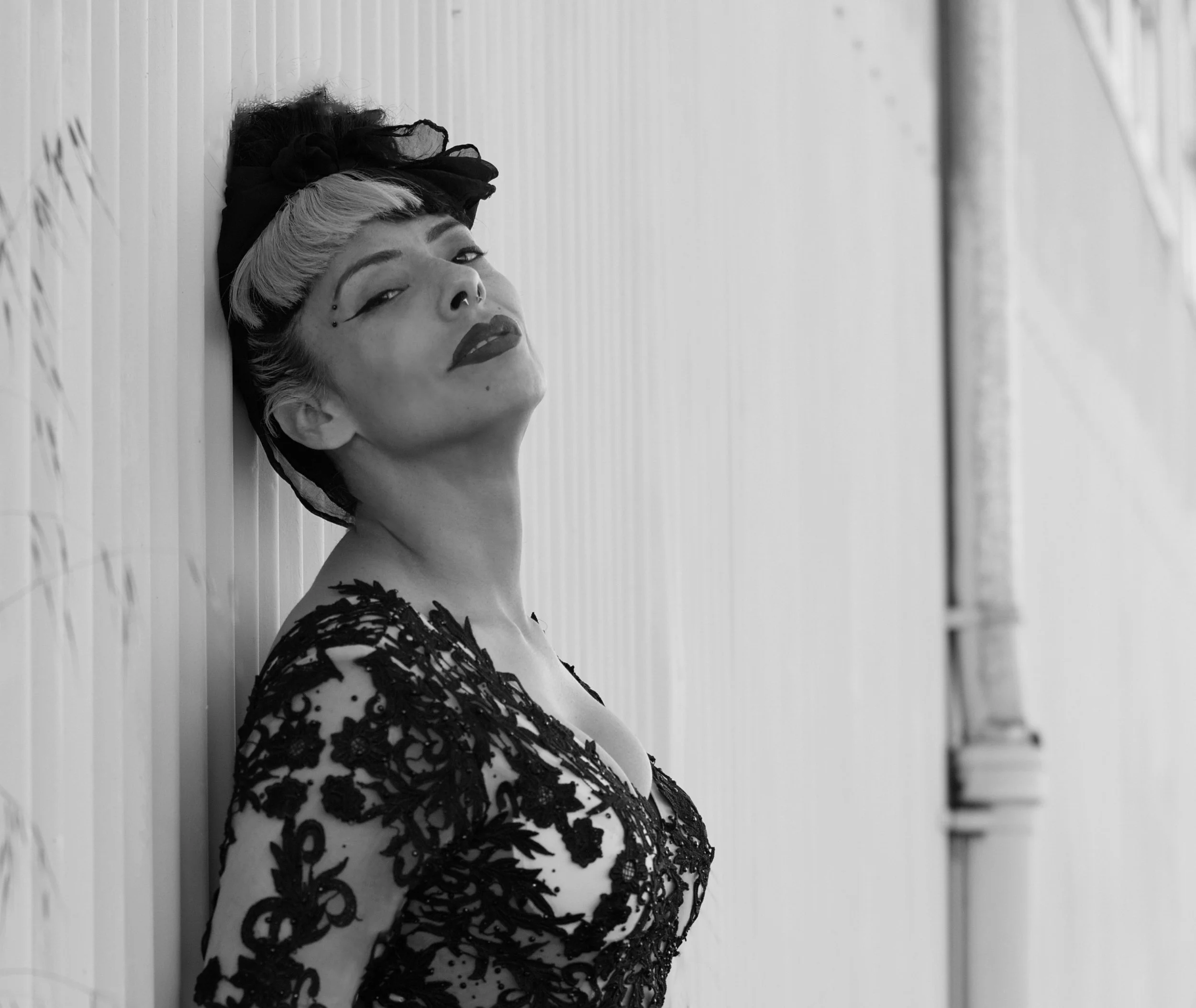Black and white photo of a woman with short highlights, wearing a black lace dress, leaning against a wooden wall, looking at the camera with a confident expression.
