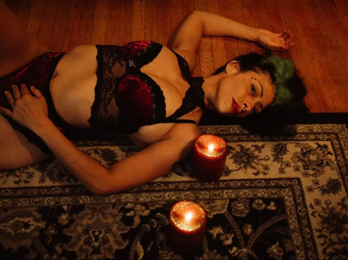 A person lying on a patterned rug with candles nearby. They have short dark hair with green highlights, are wearing black and red lace lingerie, and are smoking a cigarette.