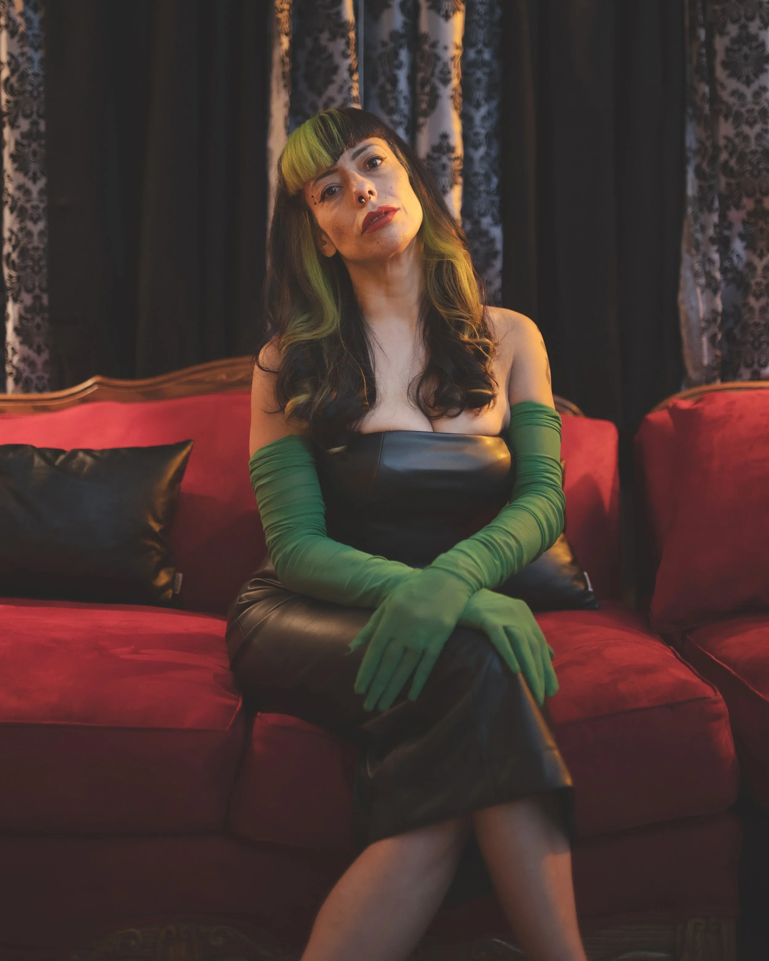 A woman with dark hair and green highlights sits on a red velvet couch, wearing long green gloves and a black leather dress, with dark patterned curtains in the background.