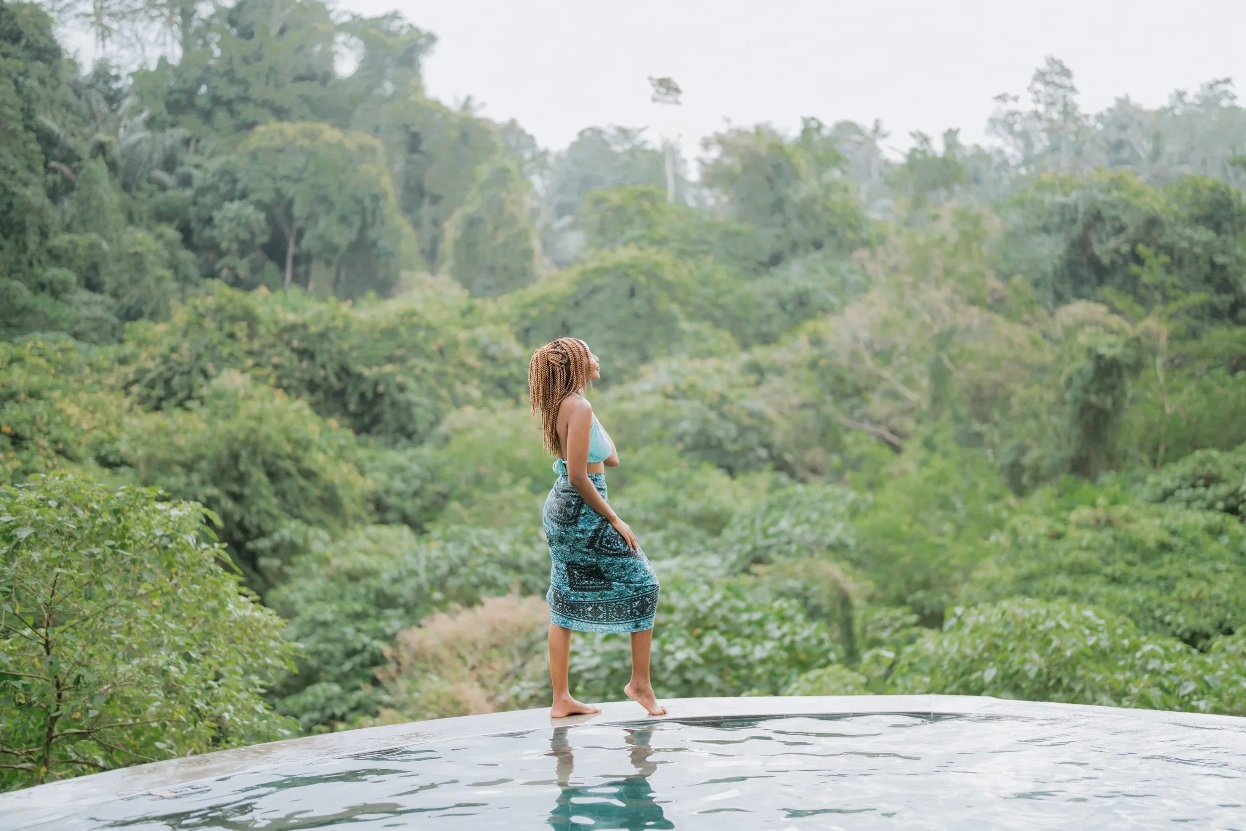 Patience Chigodora with long braided hair standing at the edge of an infinity pool overlooking a lush green jungle.