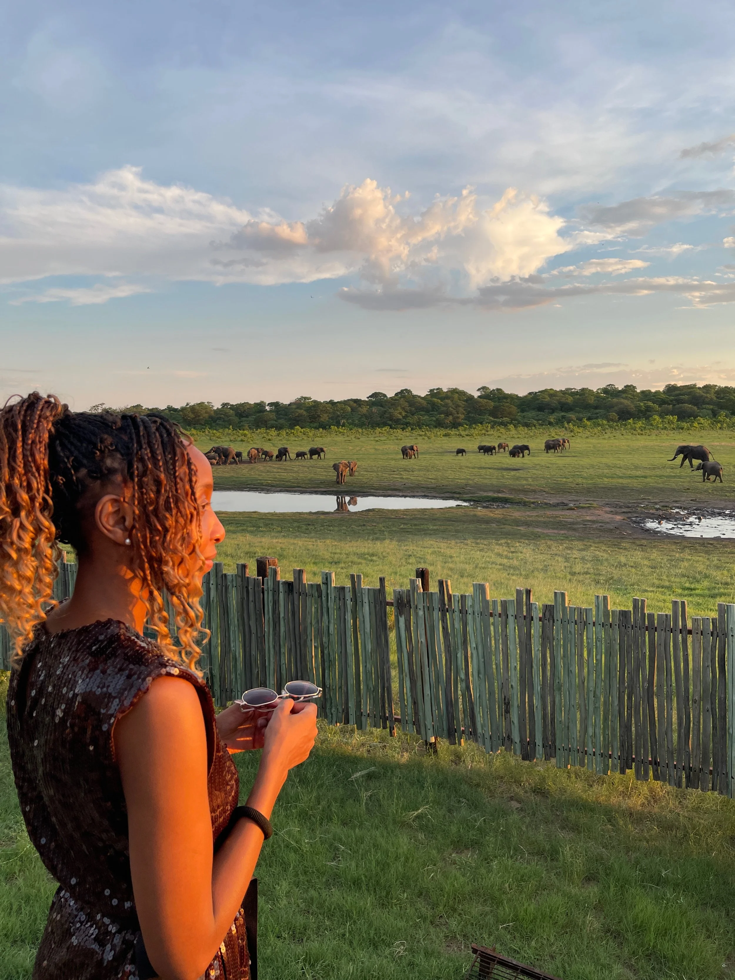 A woman relaxing in a flower-filled bathtub with a view of lush greenery outside and a statue of a woman on the side.