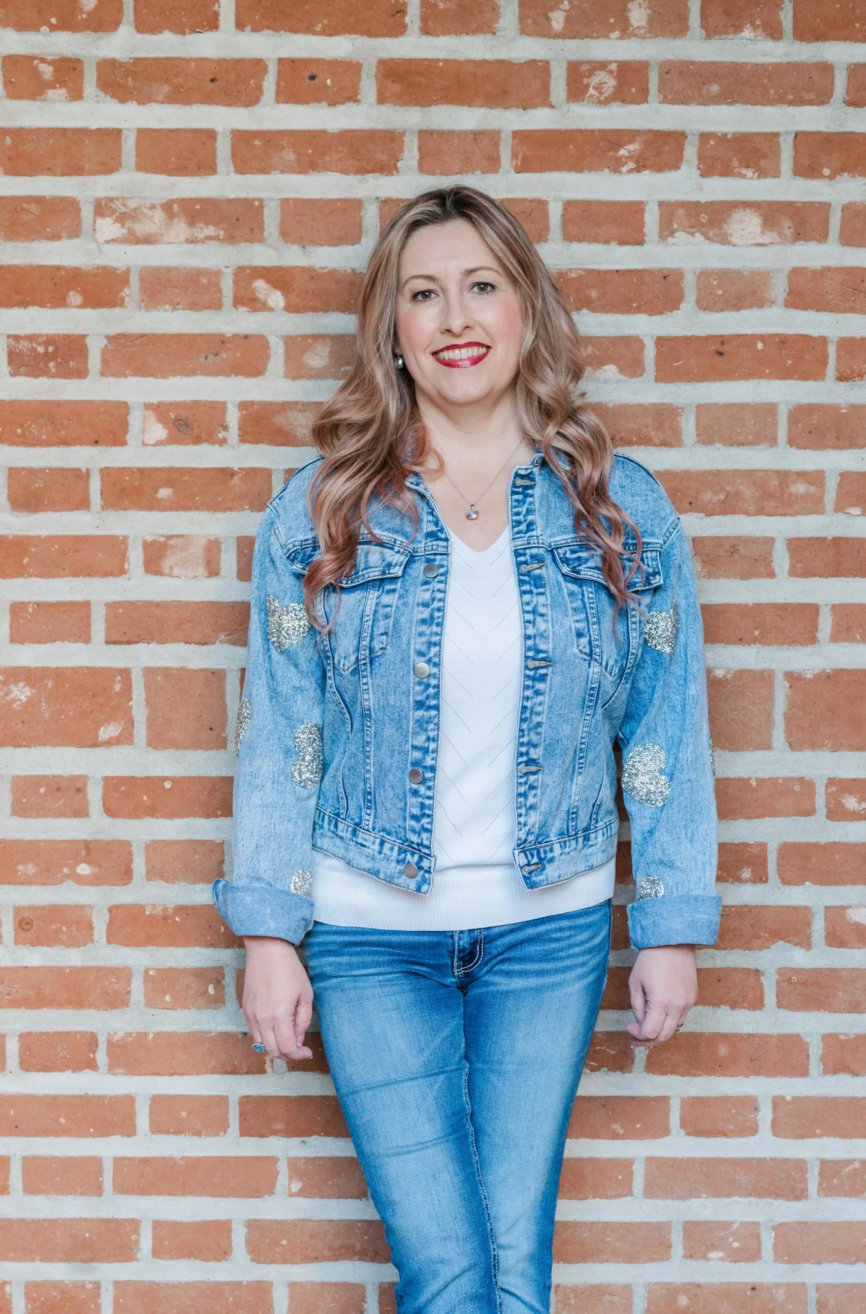A woman with long, wavy blonde hair and red lipstick, smiling while standing against a brick wall, wearing a denim jacket with silver glitter patches on the sleeves, a white shirt, and blue jeans.