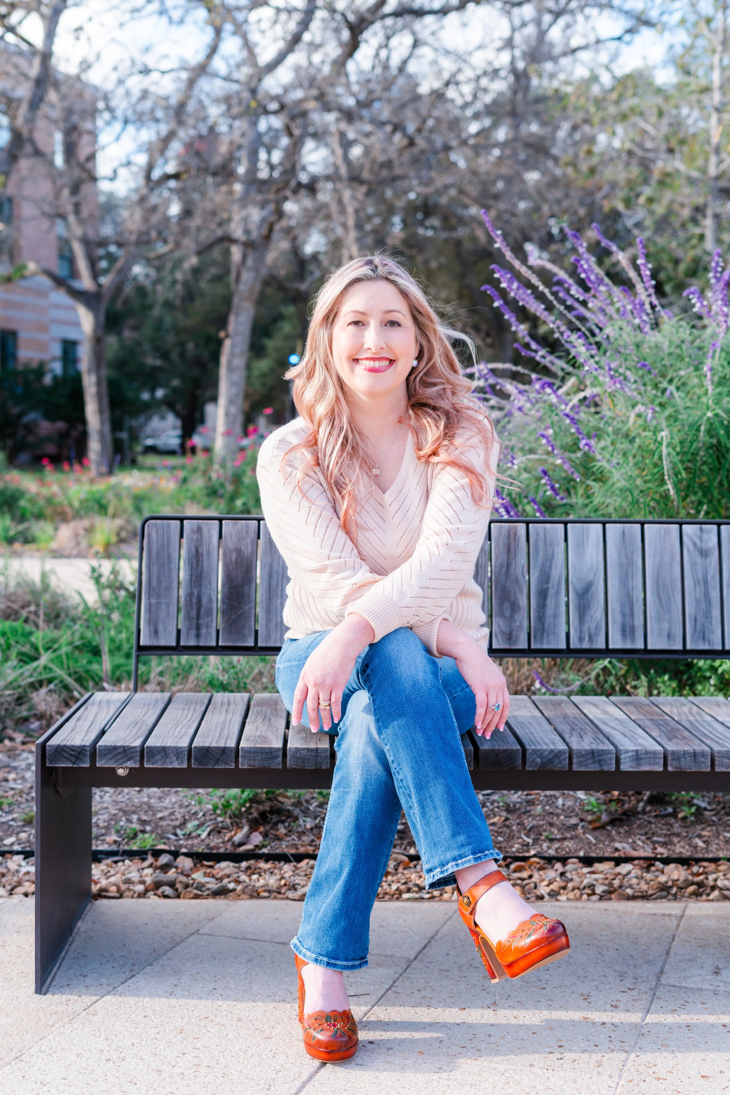 A woman sitting on a wooden park bench with her legs crossed, smiling at the camera. She has long wavy blonde hair, is wearing a cream sweater, blue jeans, and orange heels. The background features flowering plants, trees, and a building, indicating a park or garden setting during spring.