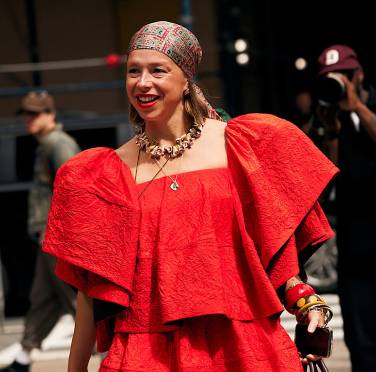Woman wearing a red textured off-shoulder dress, a colorful headscarf, and colorful jewelry, smiling outdoors at a public event.