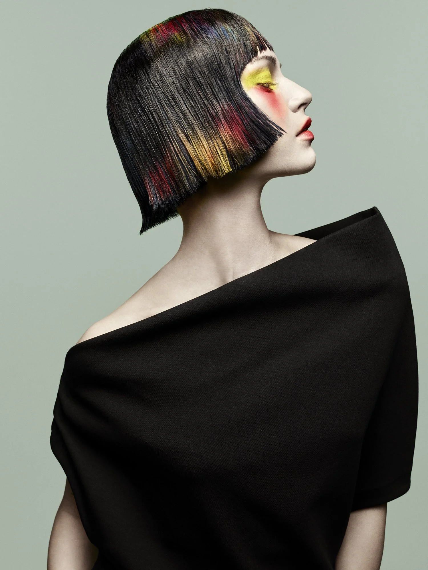 Side profile of a woman with short, straight black hair with colorful streaks, wearing a black off-shoulder top, against a plain gray background.