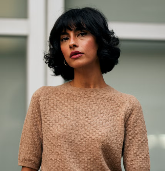 A woman with short black wavy hair and medium skin tone, wearing a beige textured top, standing indoors against a blurred background.