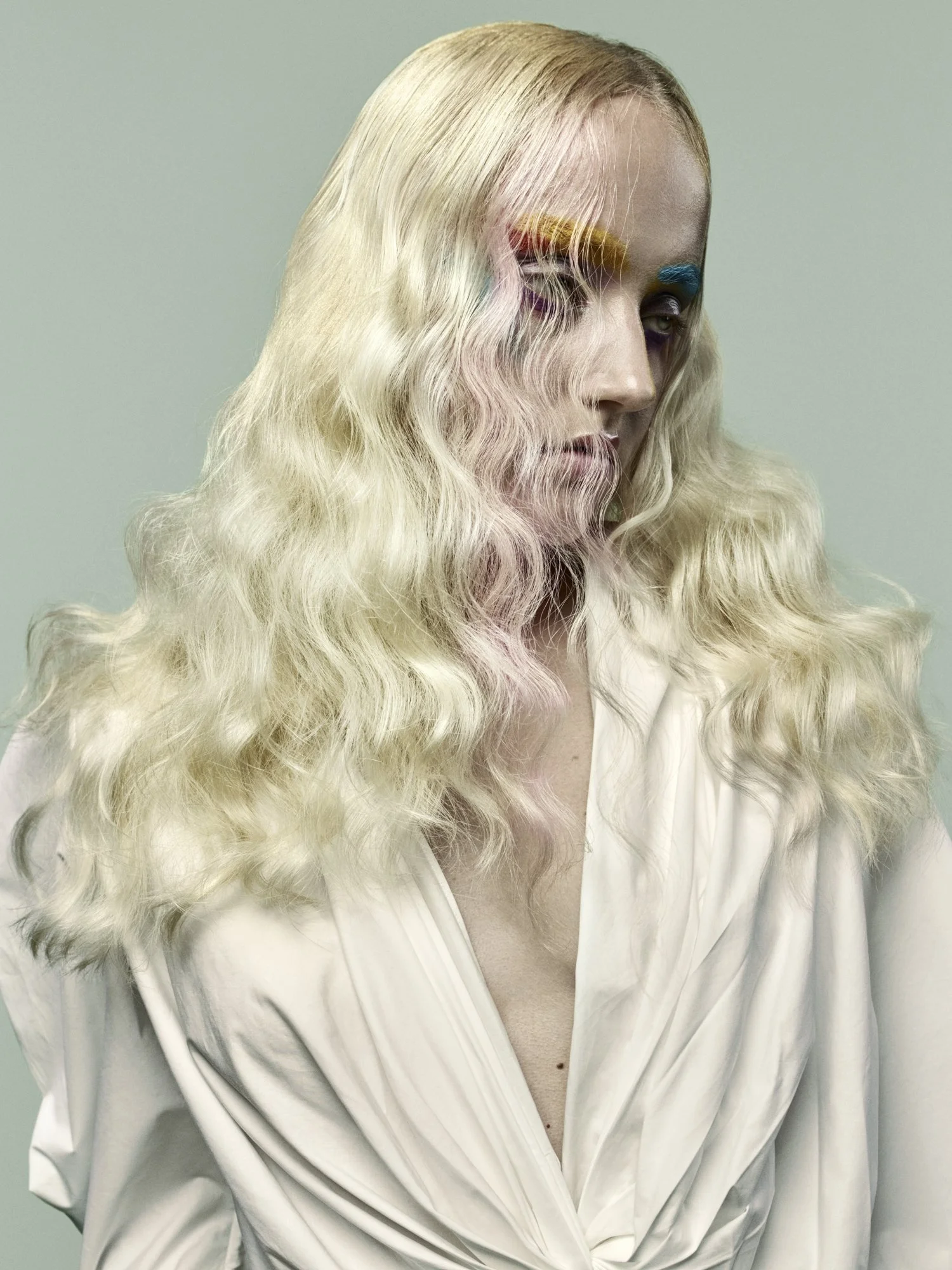 A person with long, blond, curly hair wearing a white satin shirt, with colorful makeup on their eyes and eyebrows, divided into sections of yellow, orange, purple, and blue, against a plain, light-colored background.