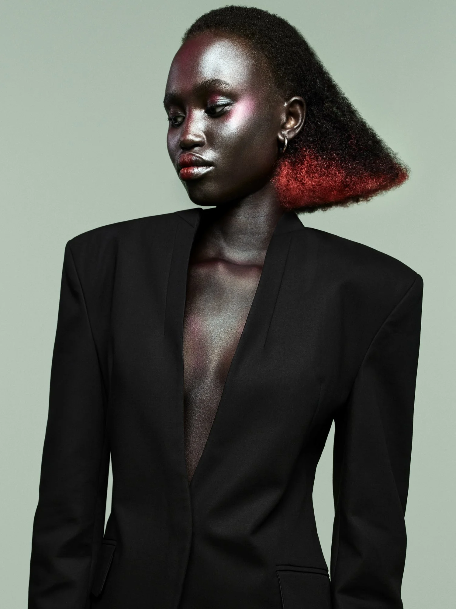 A woman with dark skin, short curly black hair. She has vibrant red and pink curly hair at the ends, wearing a black blazer with a deep V-neck, and has silver hoop earrings. She is looking down with a neutral expression against a plain light backgrou