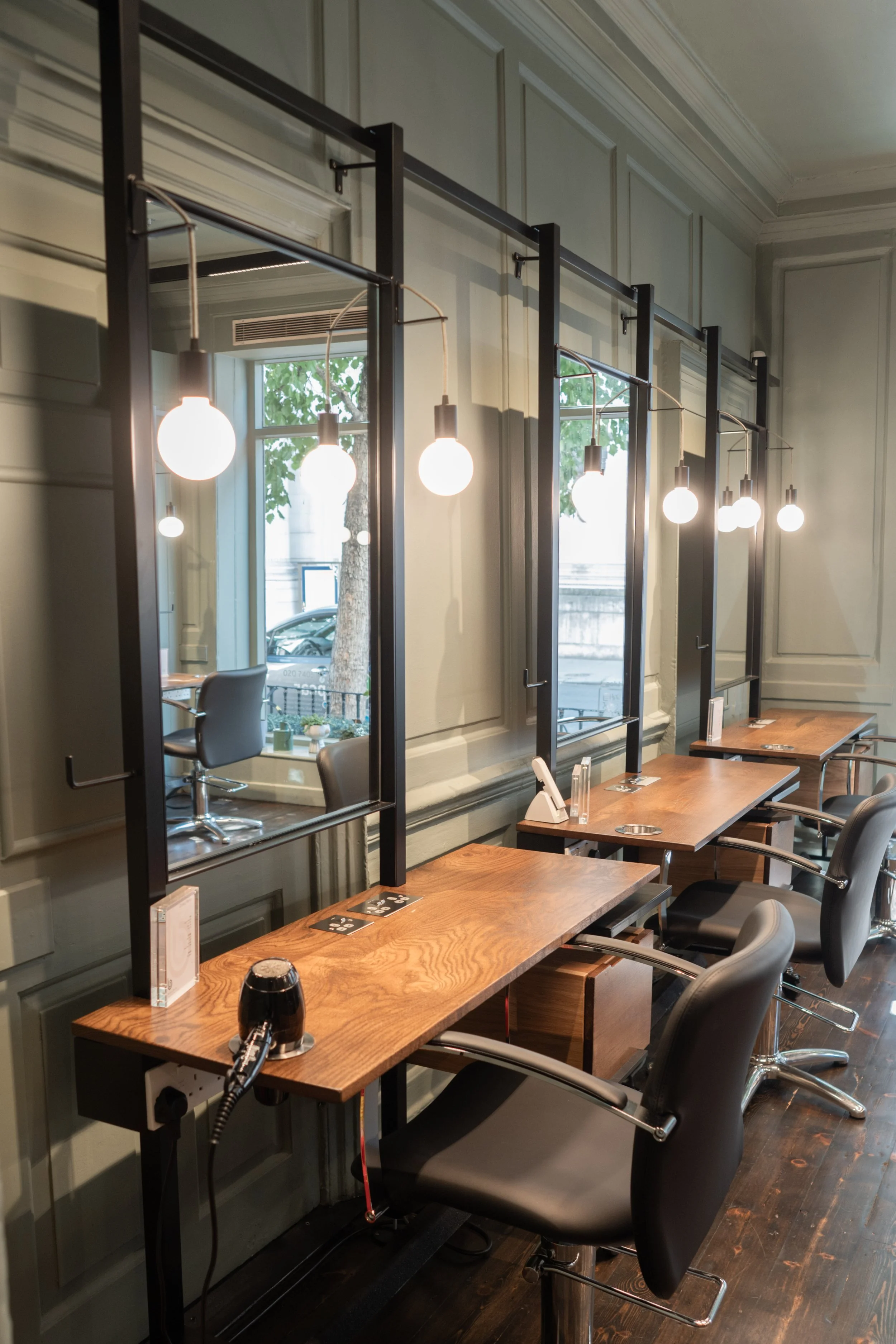 A modern salon station with three workstations, each equipped with a wooden countertop, a black chair, and adjustable mirrors with hanging light bulbs for illumination.