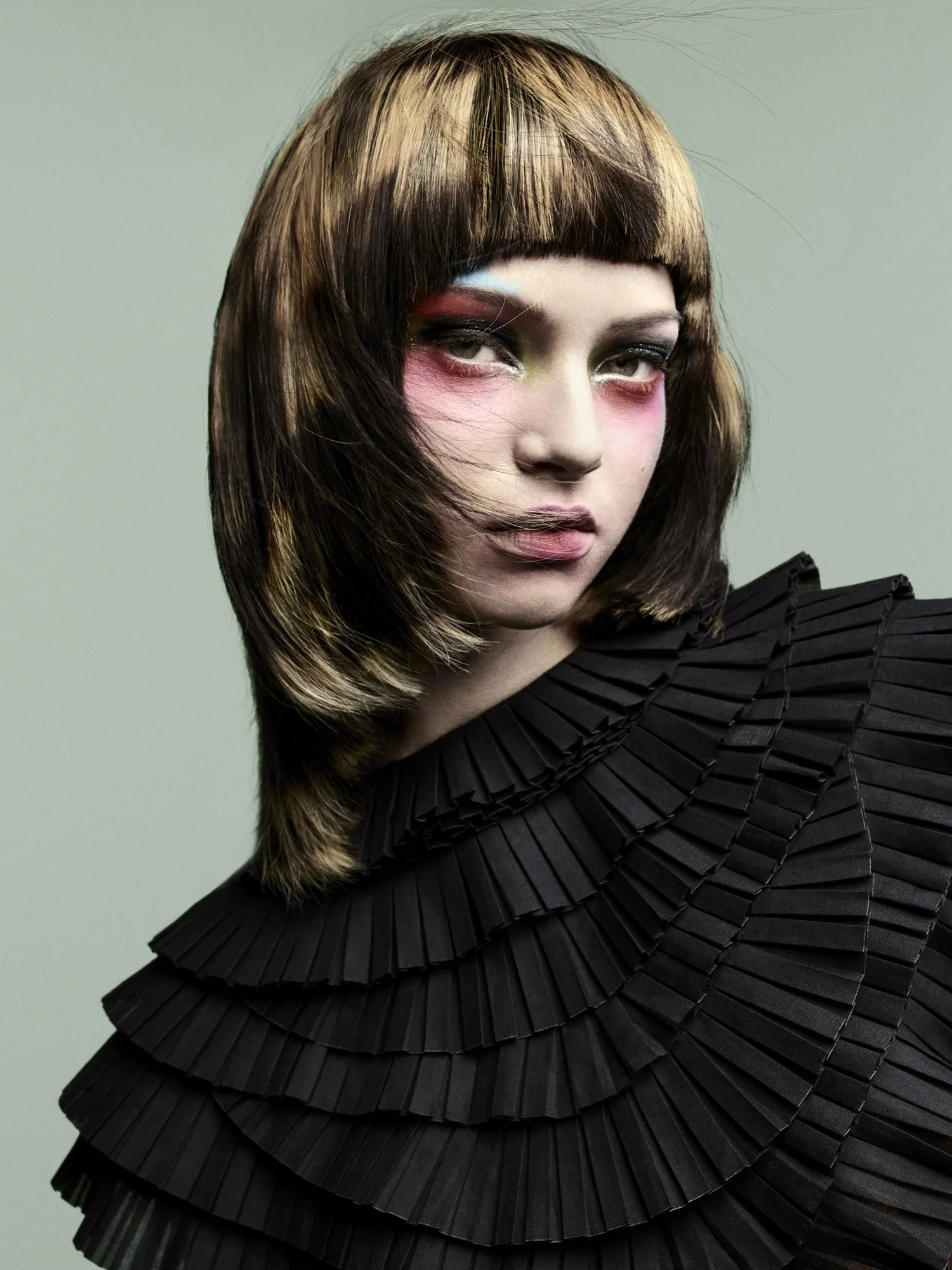 A woman with shoulder-length brown hair with blonde highlights, wearing bold colorful makeup and a black pleated garment.