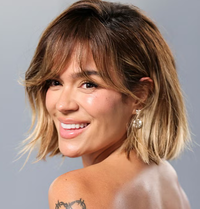 A woman with toned shoulders and a short, layered blonde haircut smiling and looking over her shoulder, wearing pearl earrings and a tattoo on her arm.