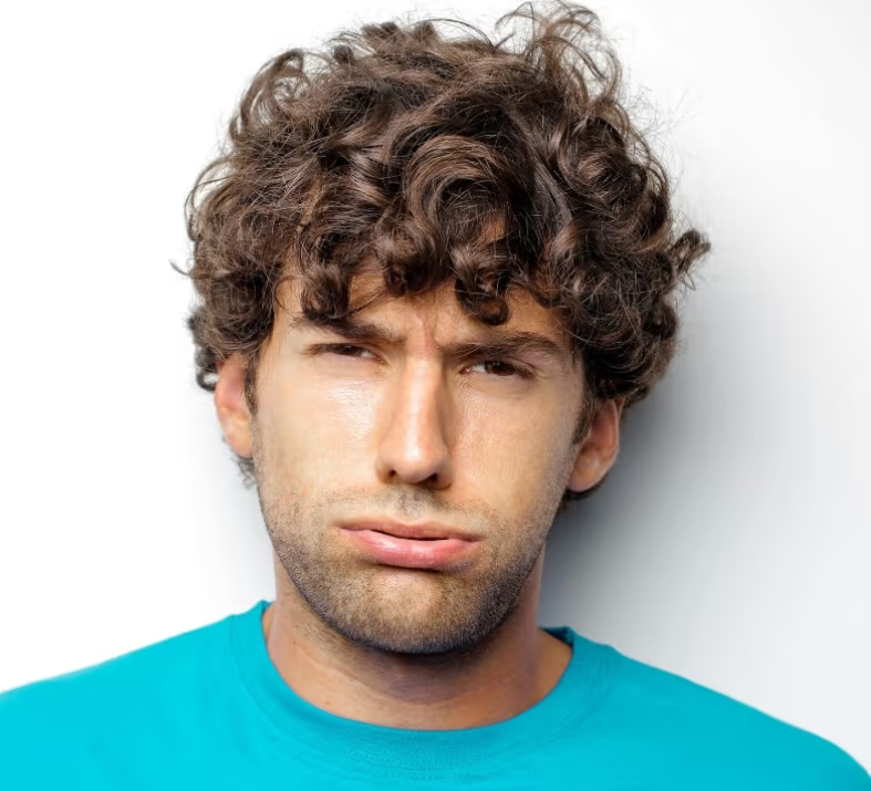 A young man with curly brown hair, wearing a blue shirt, with a confused or puzzled facial expression, standing against a white background.