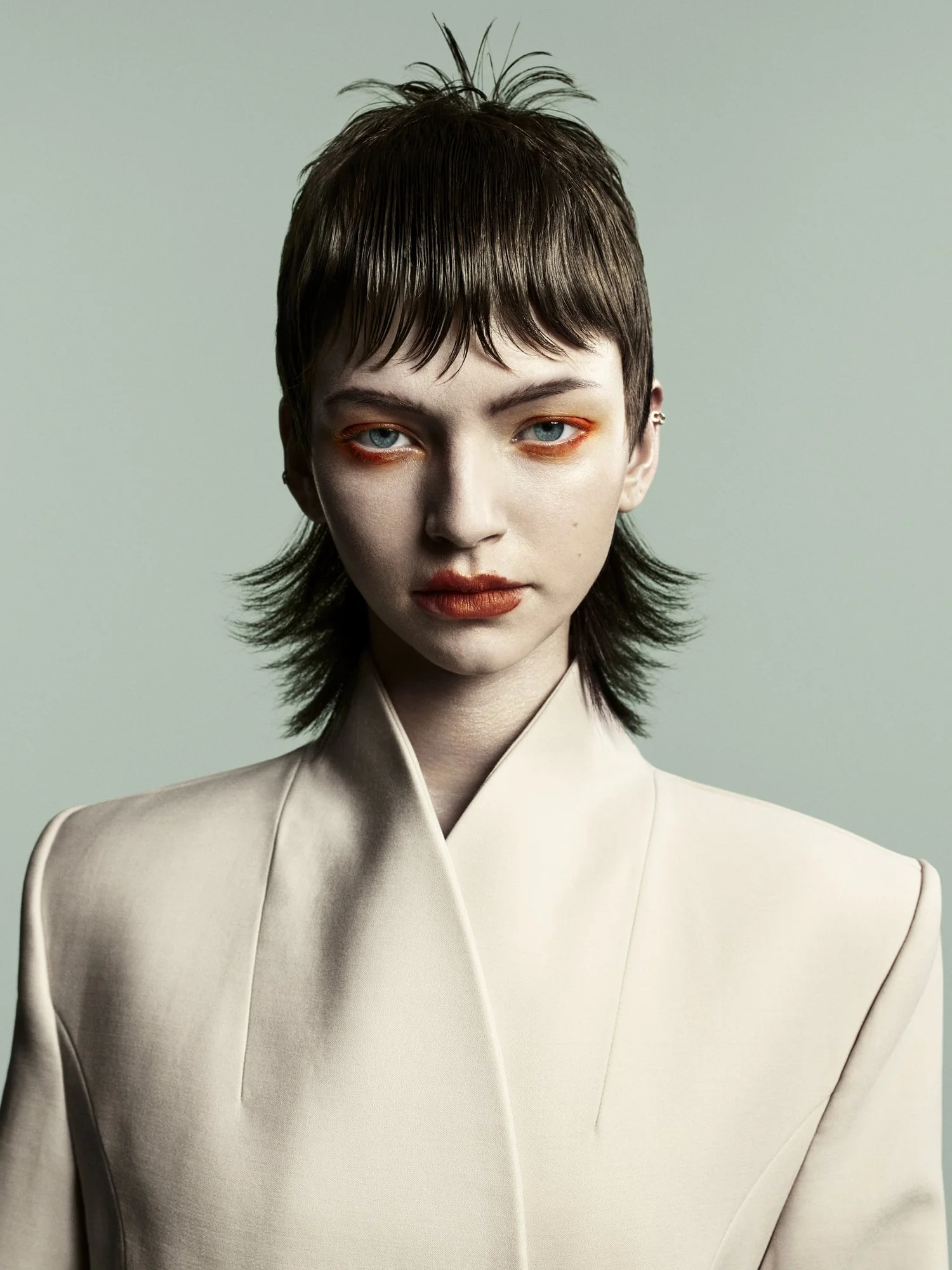 A woman with short brown hair, blue eyes, and orange makeup around her eyes and lips, wearing a cream-colored suit jacket and standing against a plain light green background.