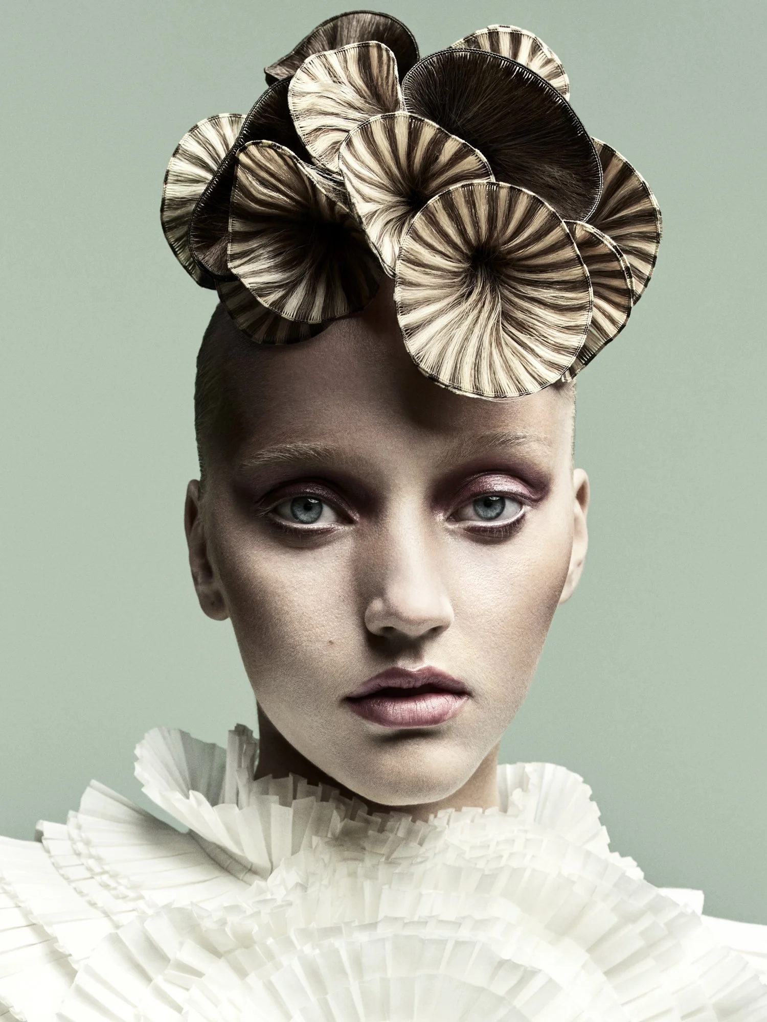 Close-up of a person with pale skin, blue eyes, and blonde hair, wearing an elaborate white ruffled collar and a headpiece made of black and beige pleated material shaped like large, overlapping fans.