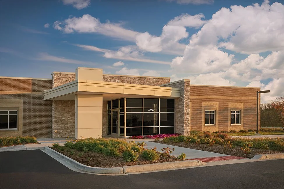 Modern commercial building with glass entrance, landscaping, and a partly cloudy sky.