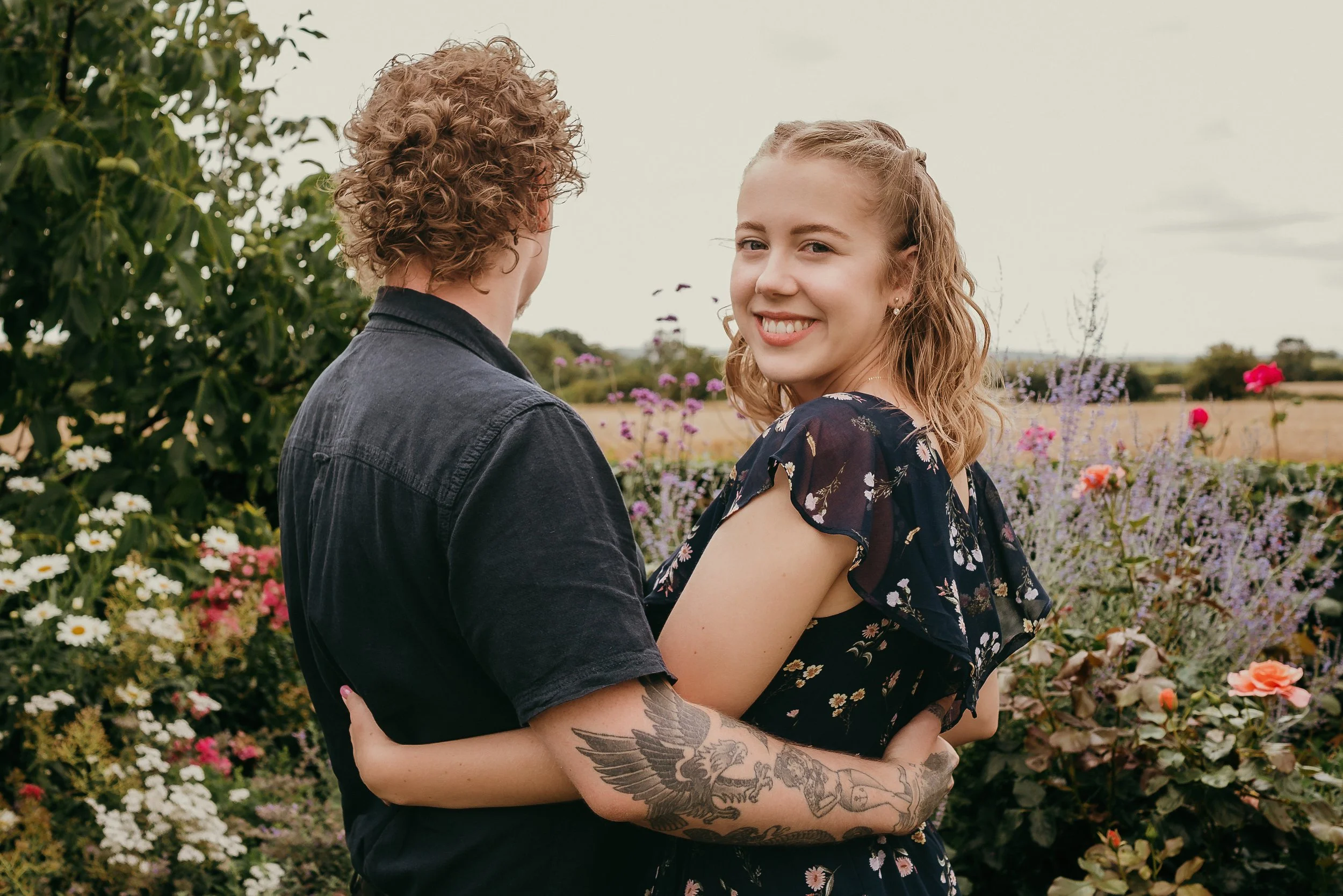A smiling woman with wavy blonde hair holds a man with curly hair in a garden of colorful flowers.
