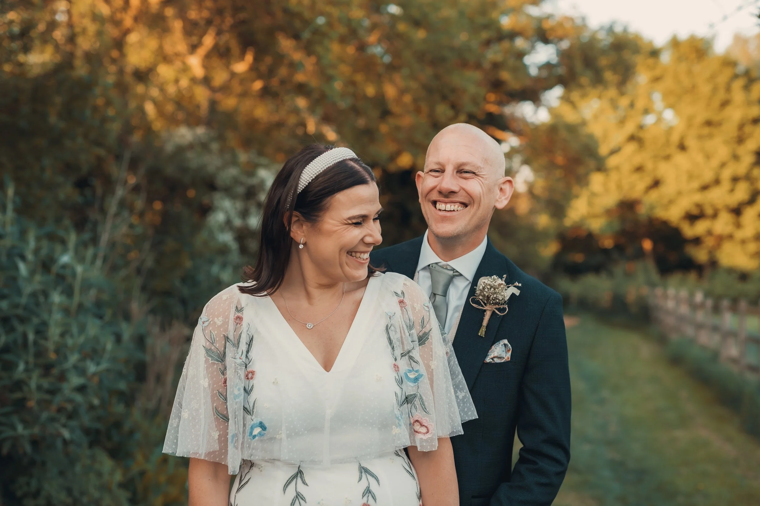 A smiling couple in wedding attire outdoors during sunset, with autumn-colored trees in the background.