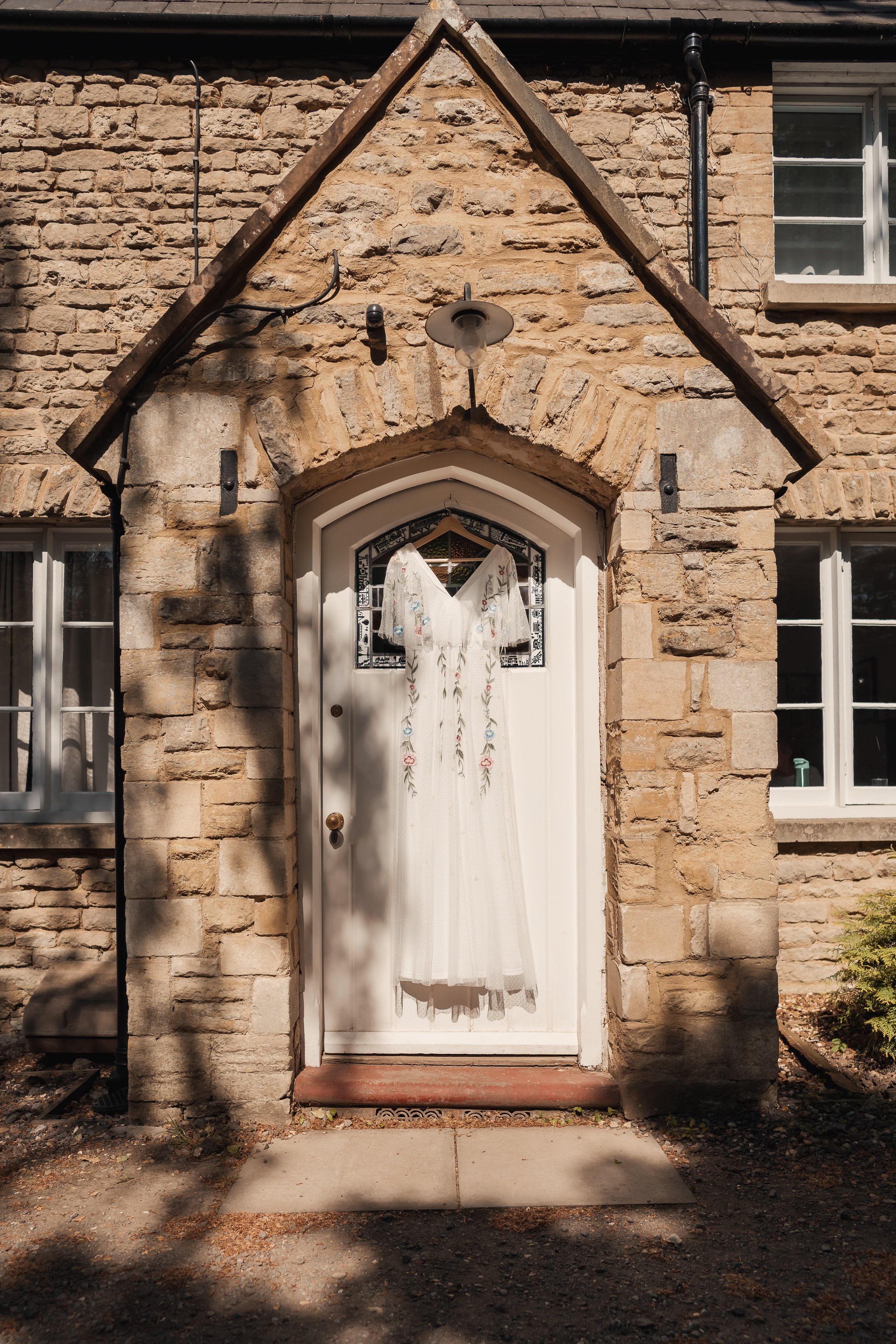 A white door with a glass window and floral embroidery hanging with a veil or dress on it. The door is set in a stone brick house with four windows visible, two on each side, with a gabled roof above the door and brick walls.