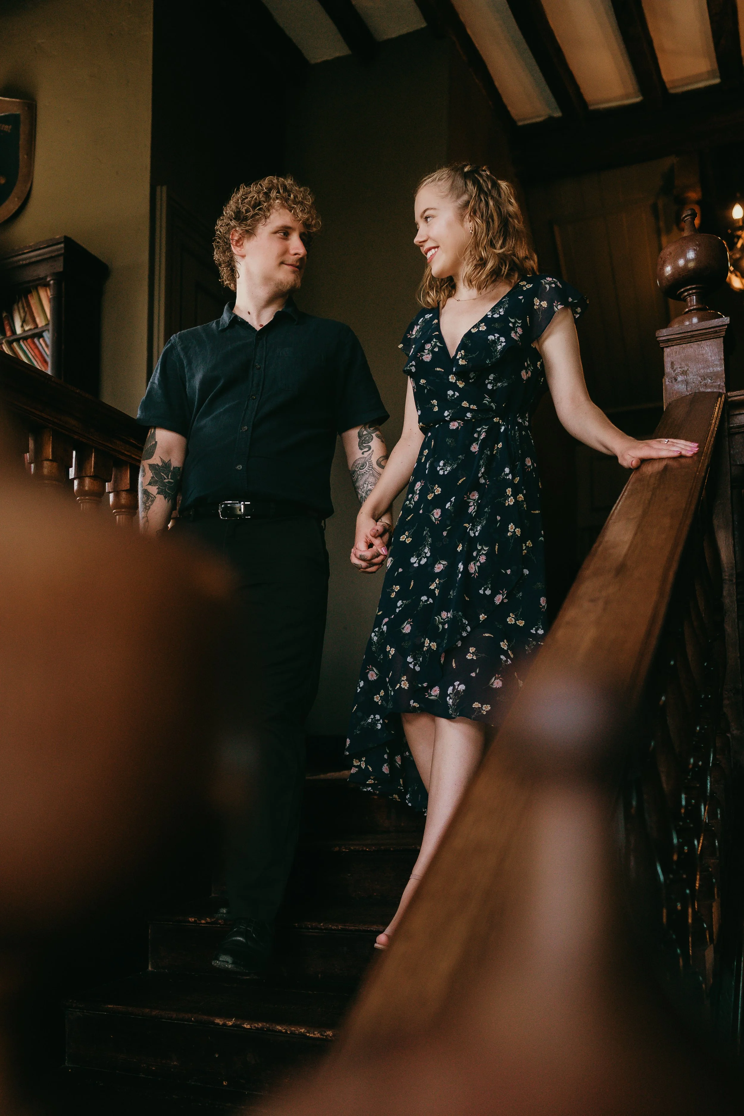A young man and woman holding hands and smiling at each other on a wooden staircase, inside a cozy, dimly-lit room with bookshelves.