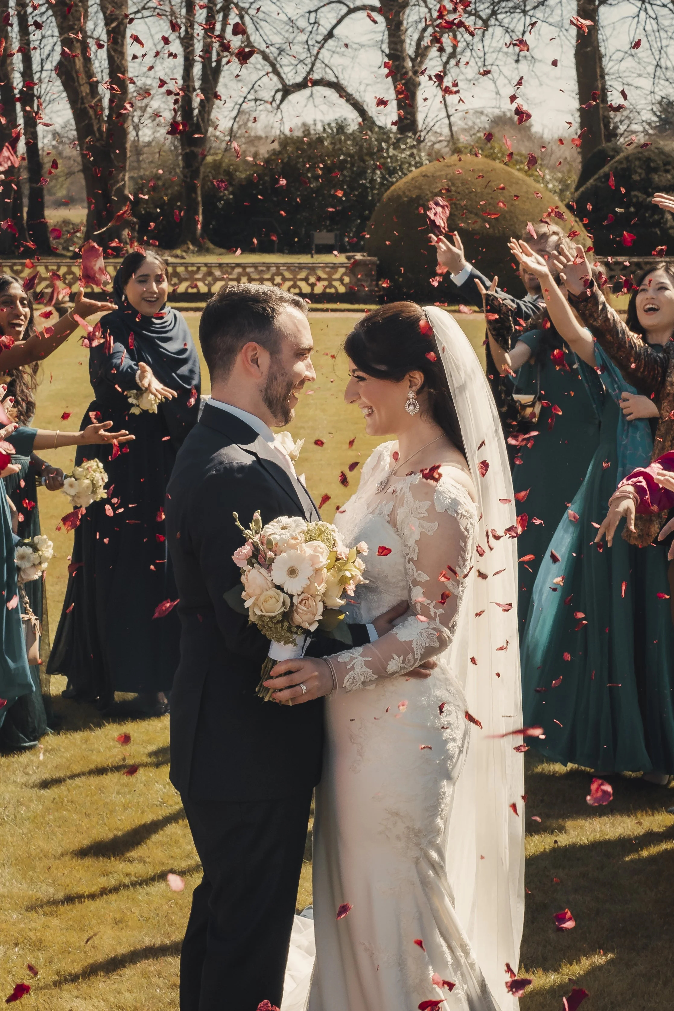 A newlywed couple sharing a kiss surrounded by friends tossing flower petals outdoors, with trees in the background.