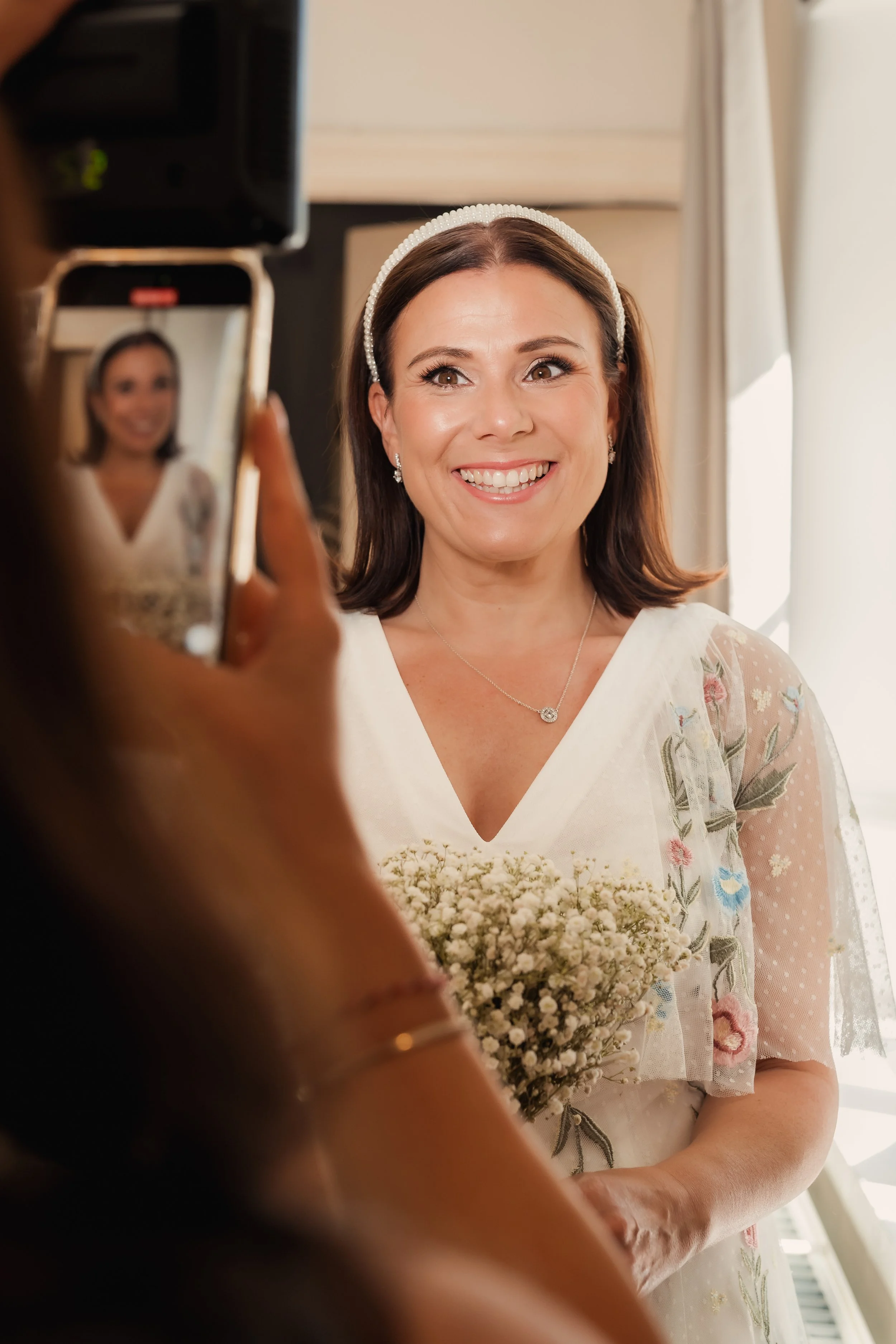 A woman in a floral dress and a pearl headband is smiling while holding a bouquet of white flowers, as someone is taking her photo with a phone.