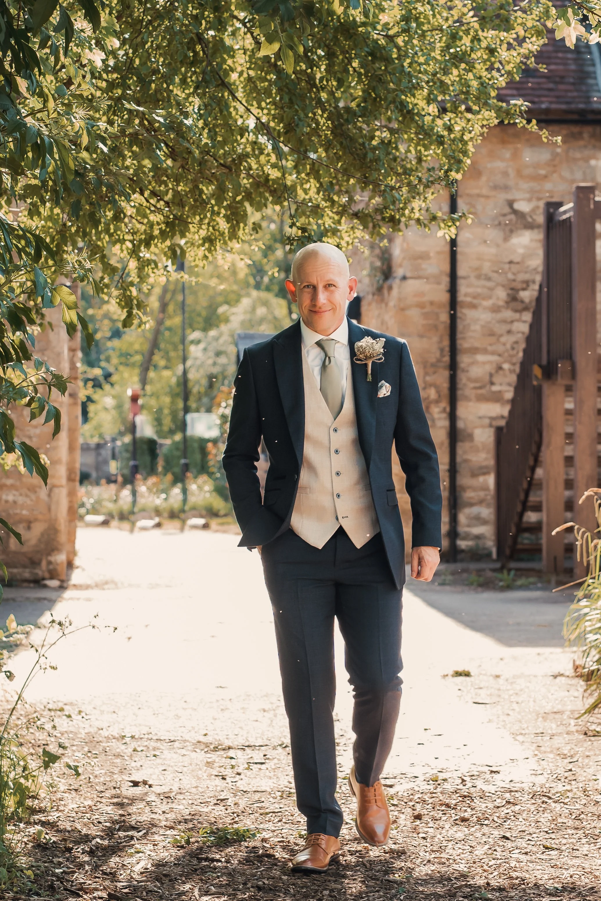 A groom dressed in a dark suit with a light-colored vest, shirt, and tie, walking outdoors under leafy trees, with a rustic stone building in the background.