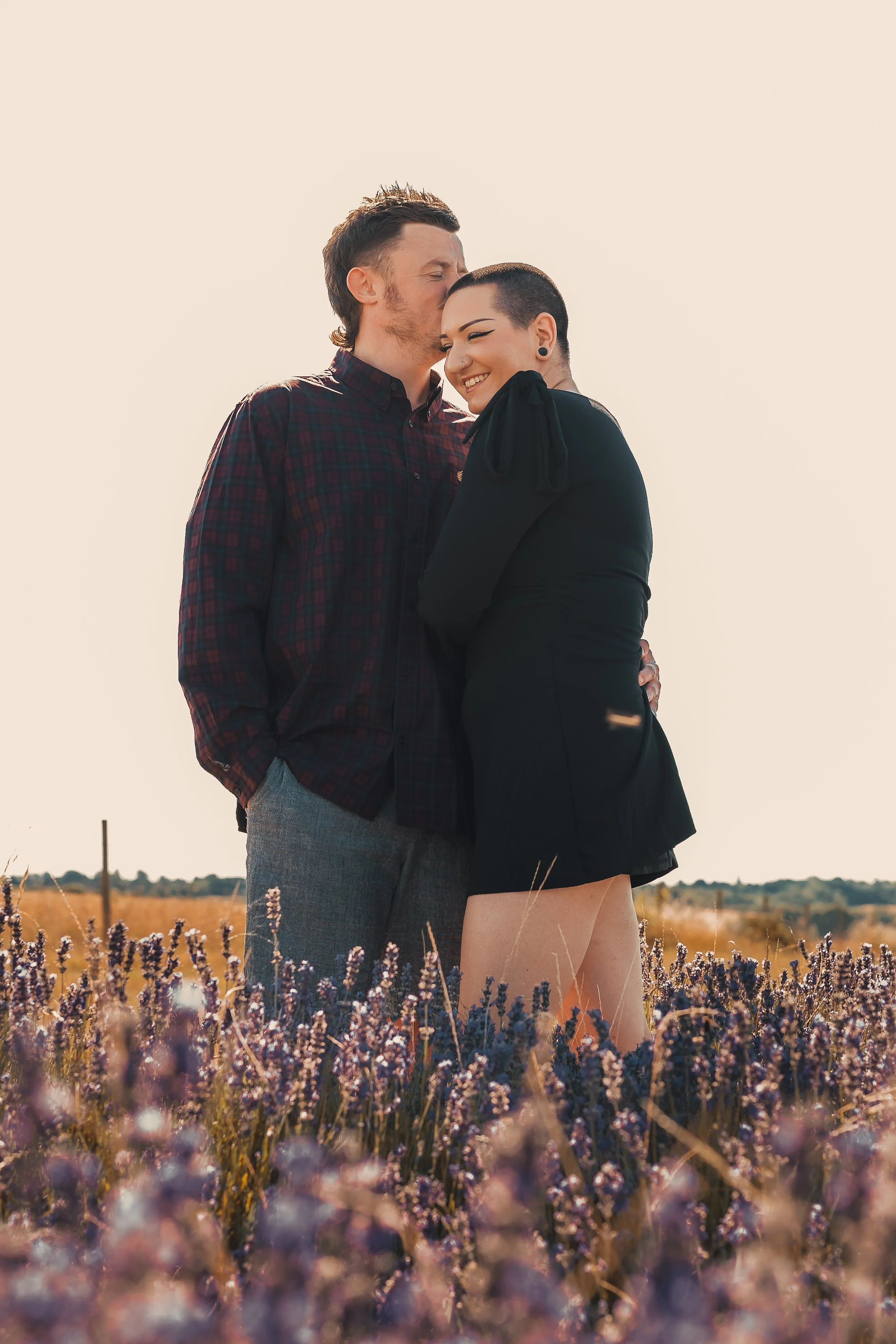 A couple standing in a lavender field during sunset, with the man kissing the woman's forehead and the woman smiling with her eyes closed.