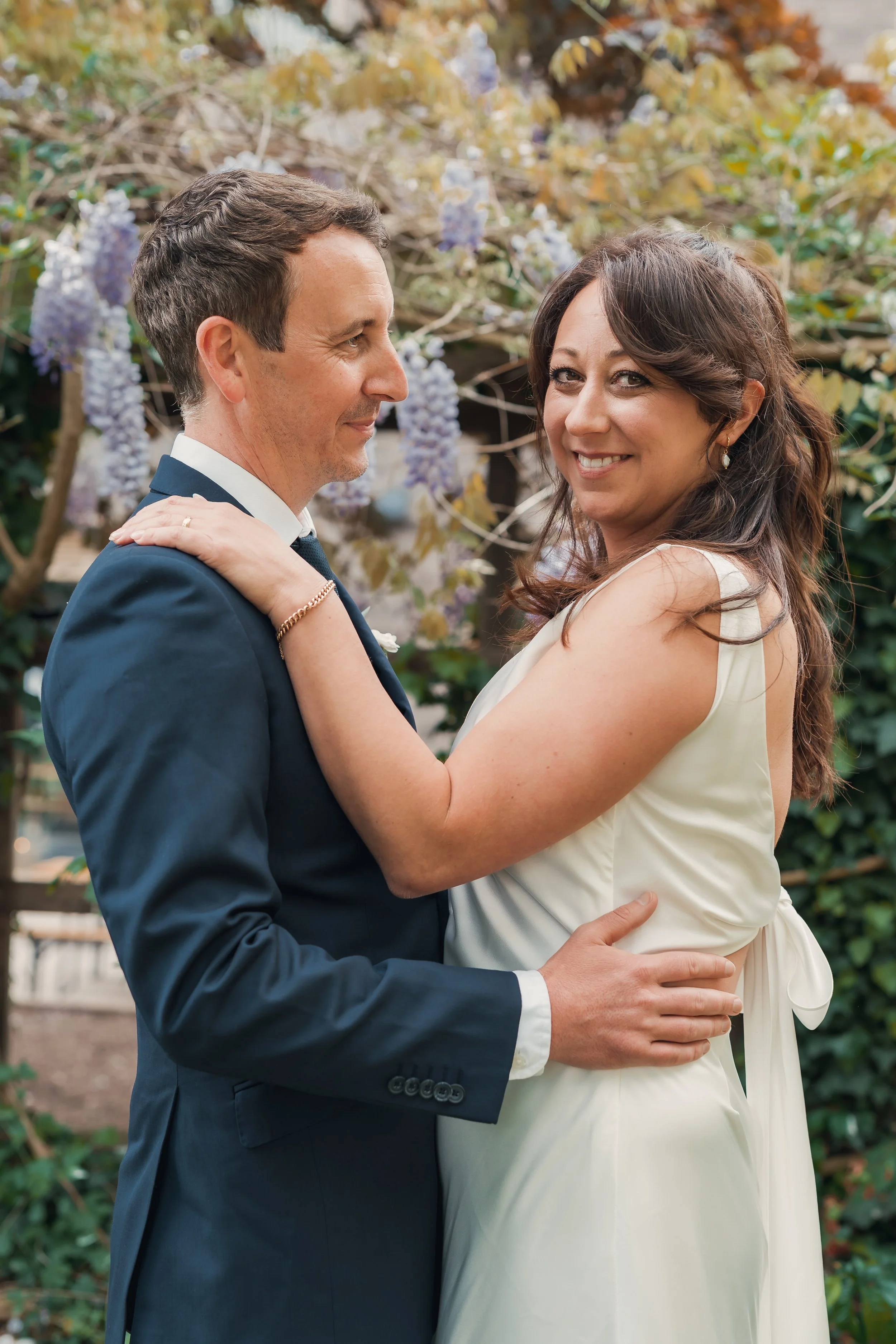 A couple dressed in wedding attire embracing outdoors, with flowers and greenery in the background.