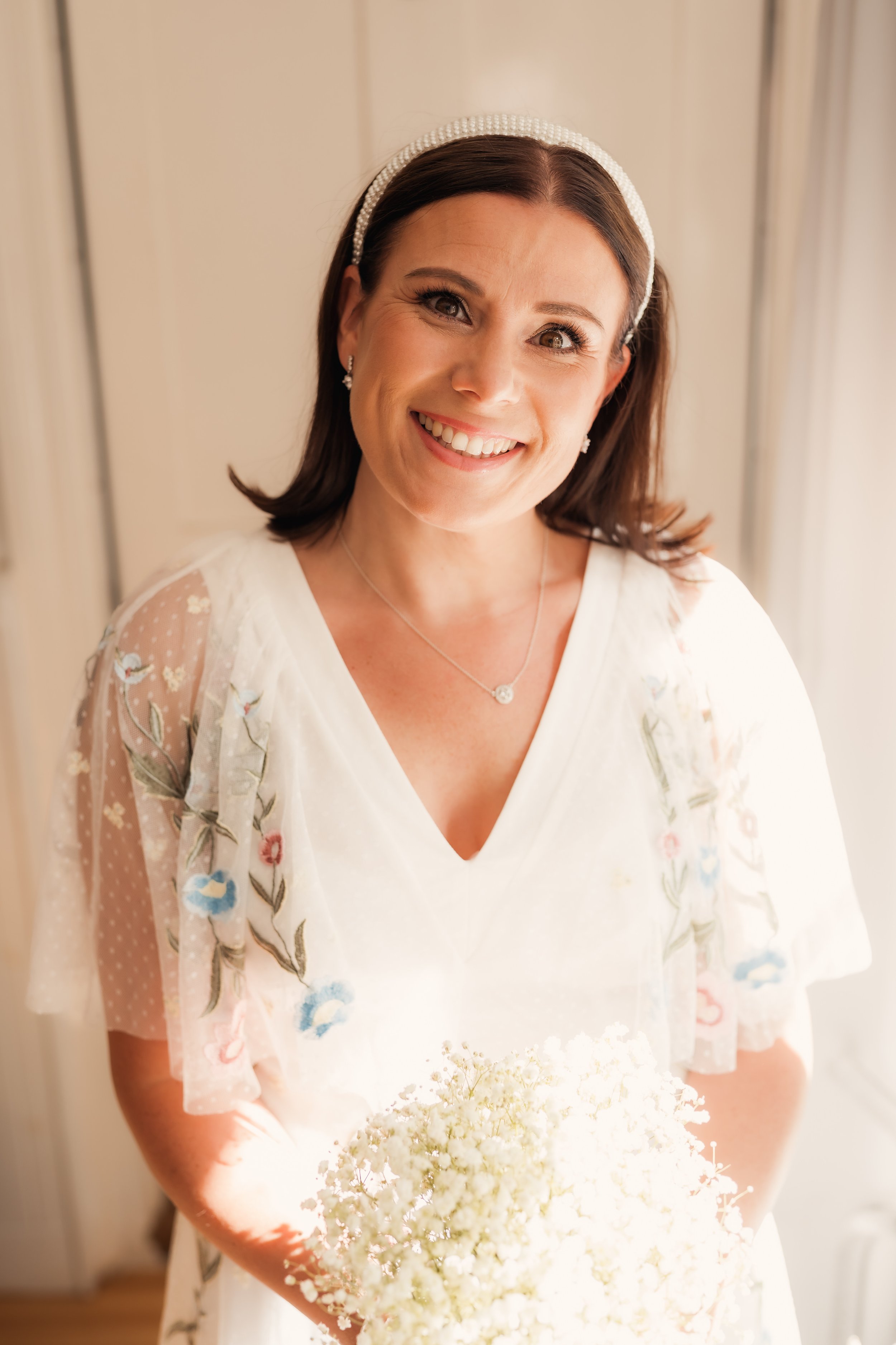 A woman with dark brown hair wearing a white dress with floral embroidery, smiling, holding a bouquet of white flowers, standing near a window with soft lighting.