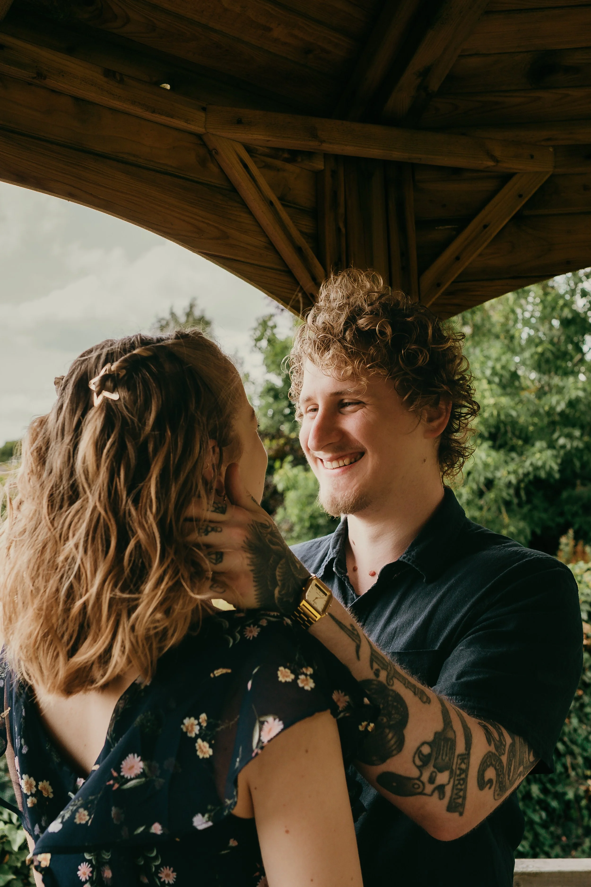 A young man and woman are sharing an intimate moment under a wooden roof. The man is smiling warmly at the woman, gently holding her face with his hand. The woman has wavy, shoulder-length hair and is wearing a floral dress. The man has curly hair, a