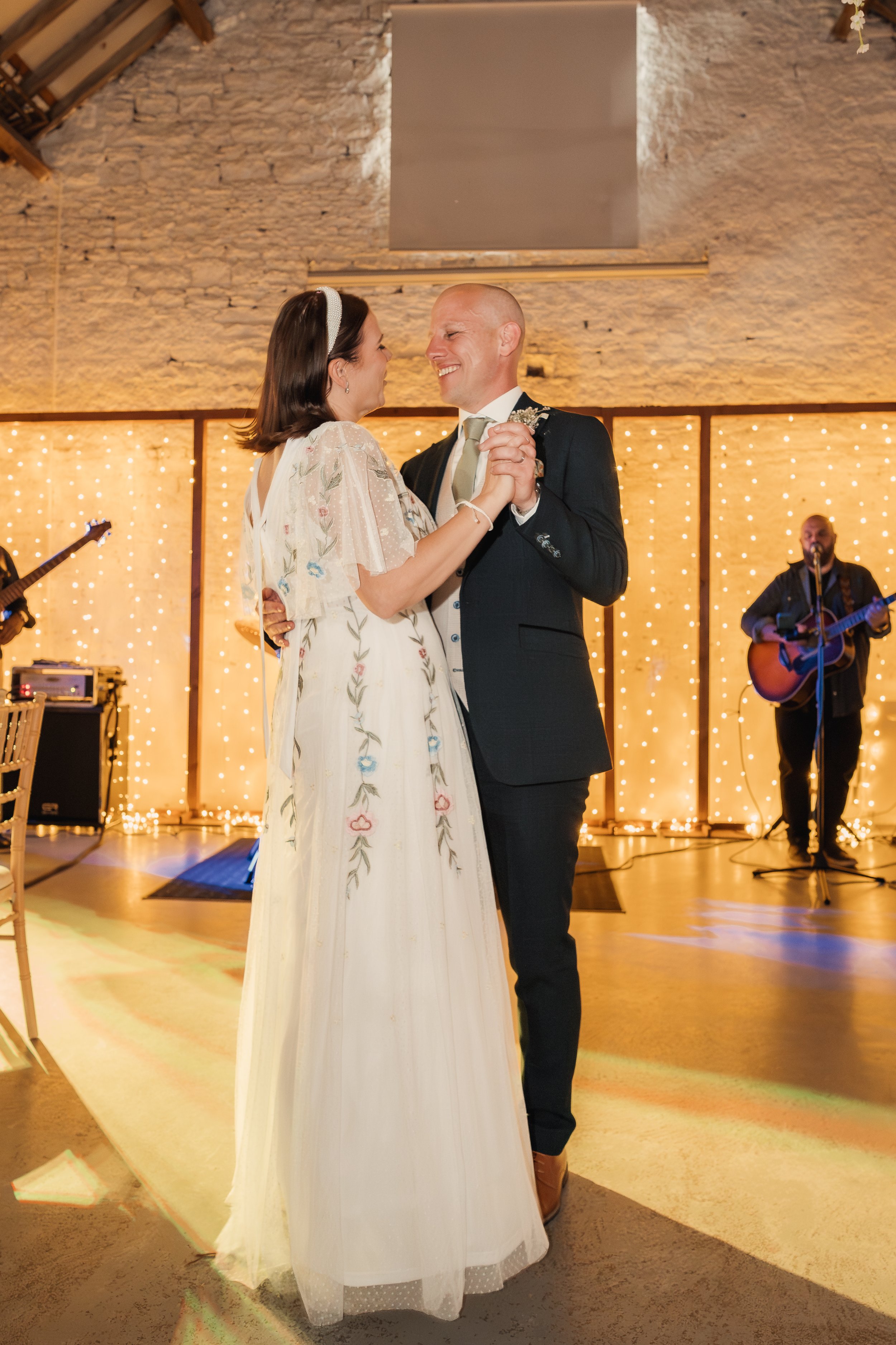 A bride and groom dancing together at their wedding reception, smiling and holding hands, with a live band playing in the background and warm string lights creating a cozy atmosphere.